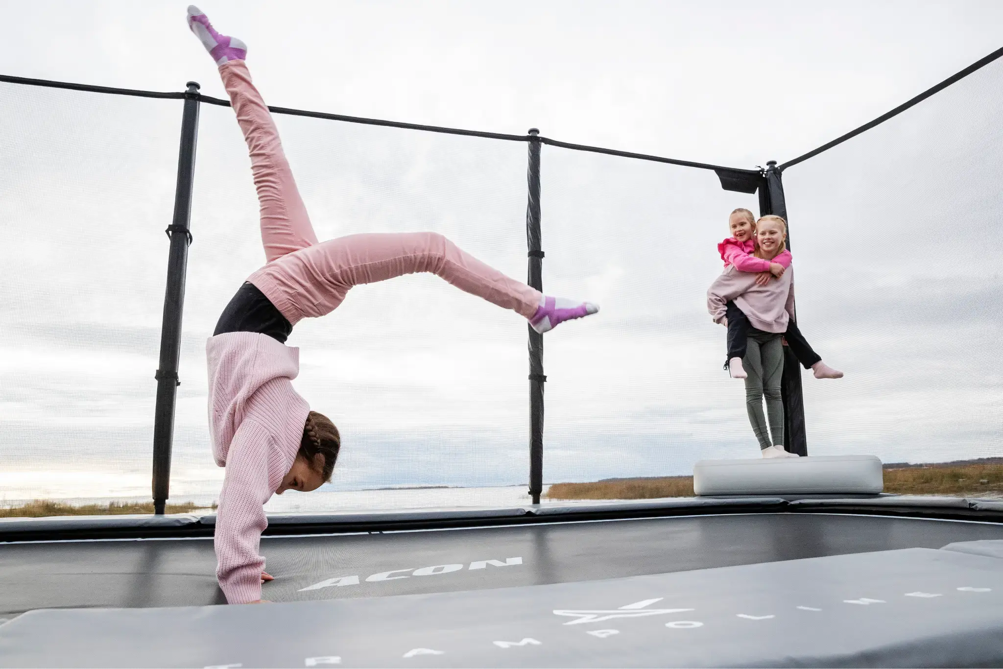 Children playing on an Acon trampoline by a lakeside. One child performs a handstand while another carries a younger sibling on their back, surrounded by a safety net.