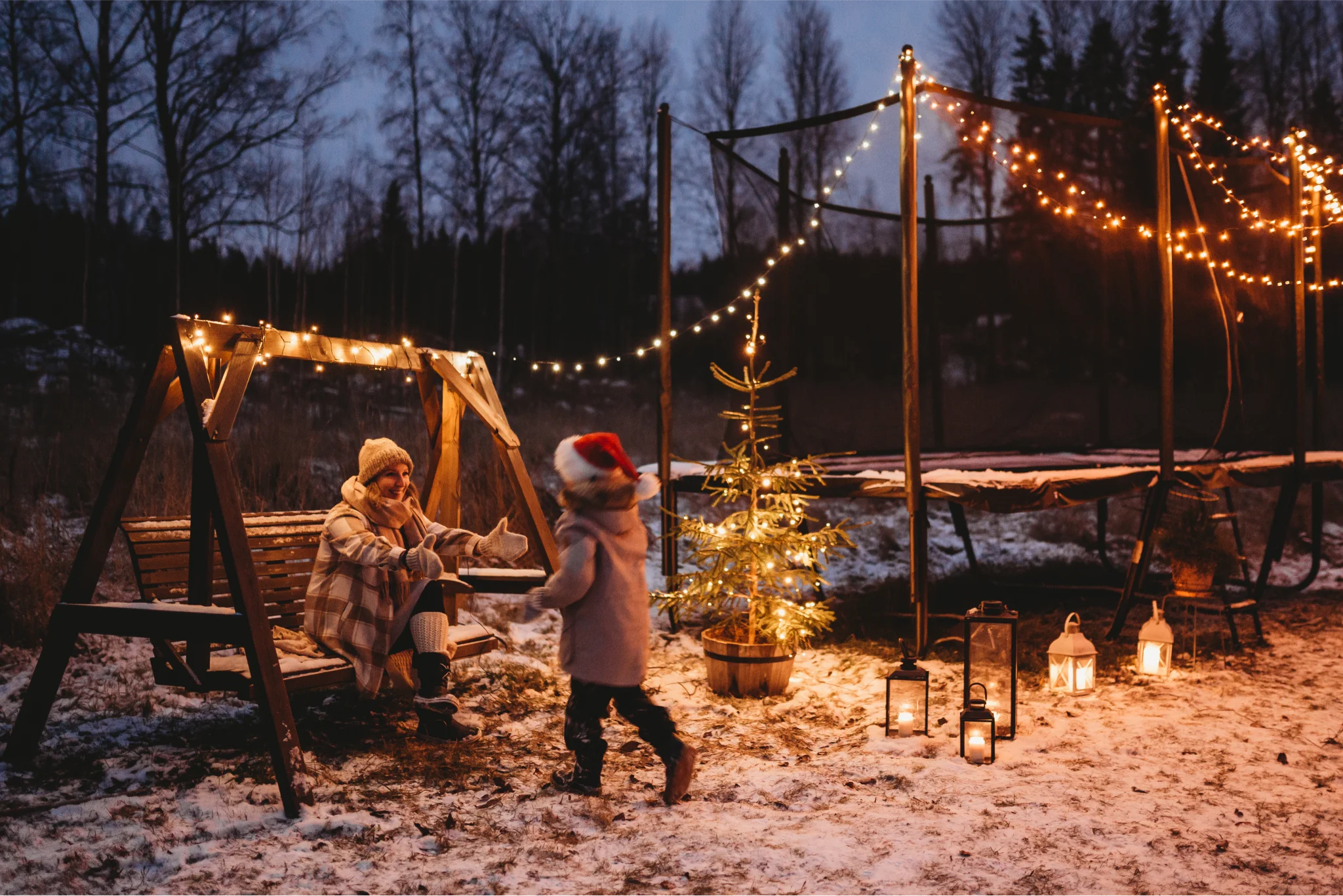 Child with a santa hat running to a woman on a swing. Snow on the ground and a christmas tree and a trampoline in the background, both decorated with christmas lights.