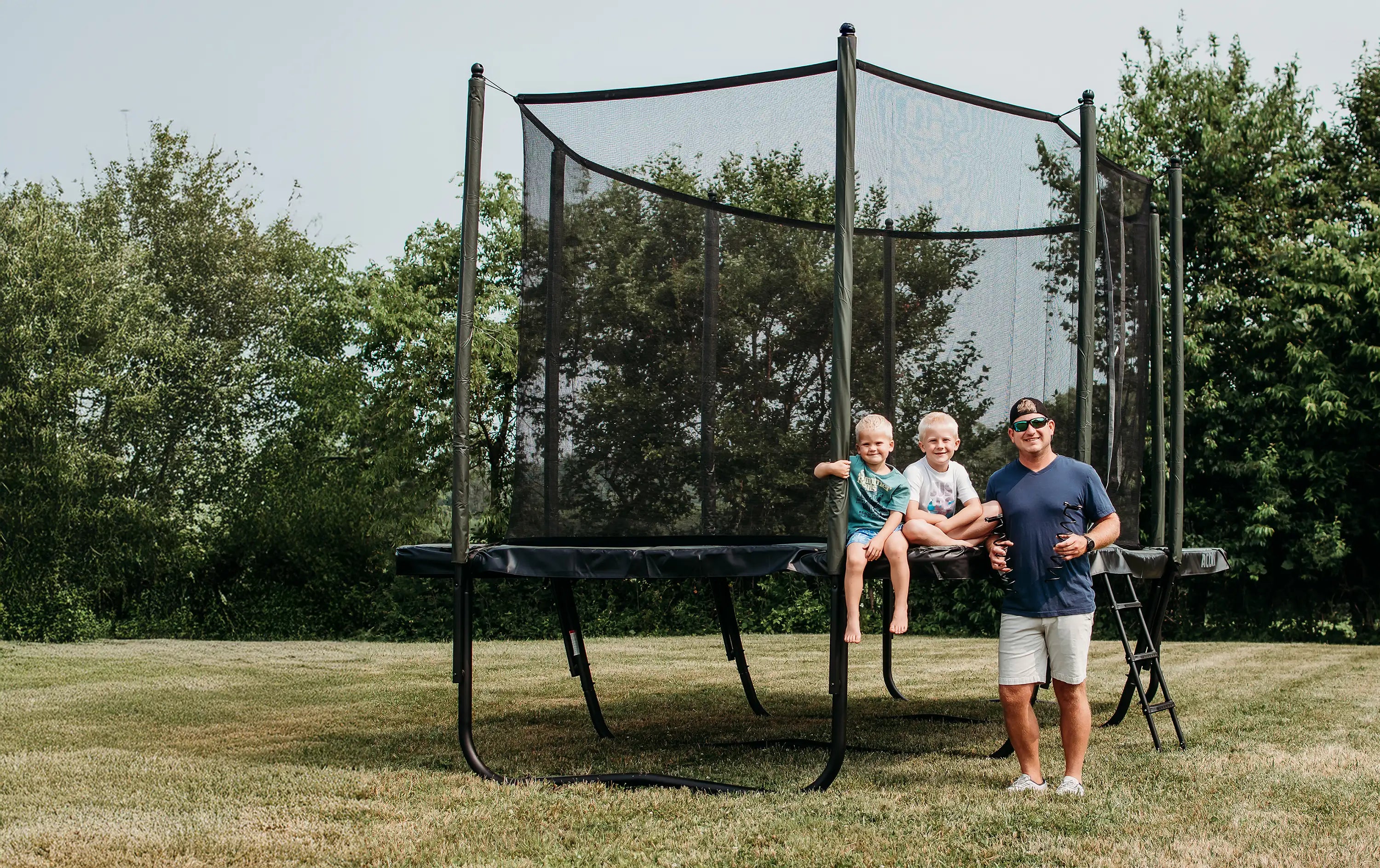 Man and two children on a Acon HD trampoline in a backyard with trees in the background.