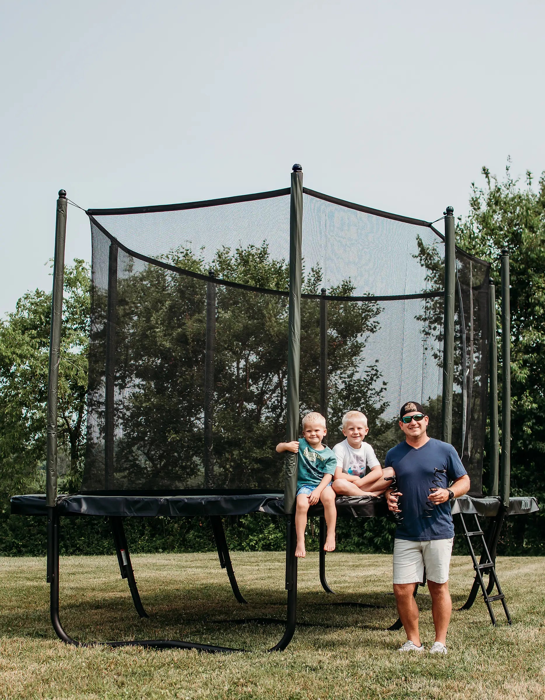 Man and two children on a Acon HD trampoline in a backyard with trees in the background.