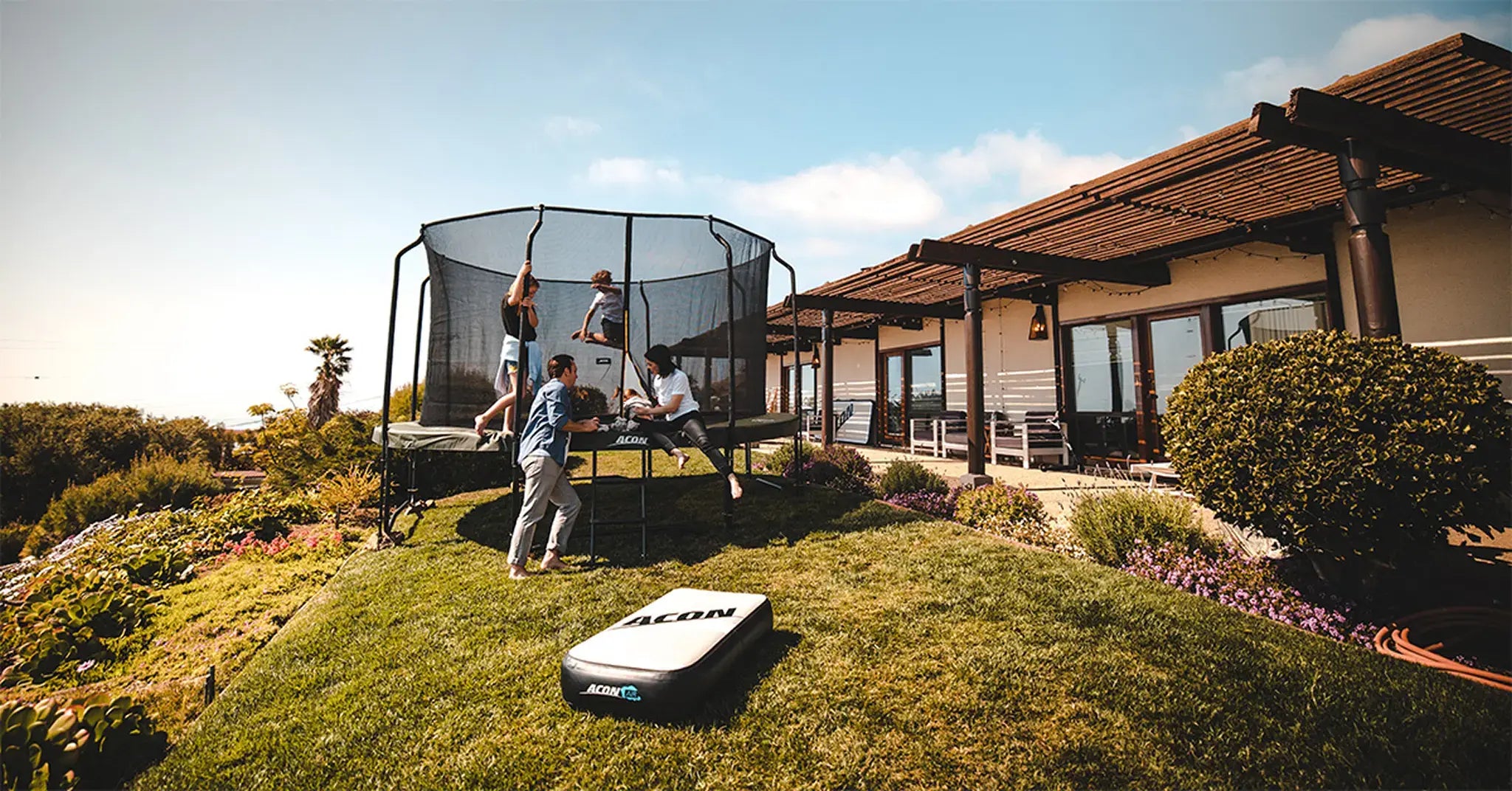 Family enjoying an Acon trampoline in a sunny backyard next to a modern house.