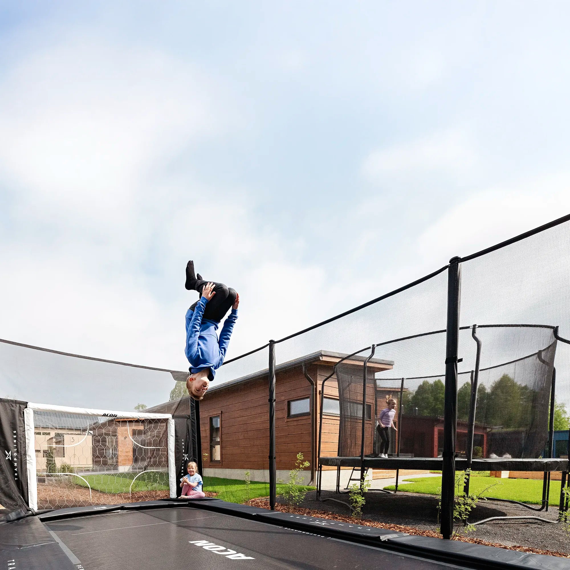 Boy flips on Acon X trampoline, little girl claps. In the neighbor’s yard, a girl jumps on Acon Air trampoline.