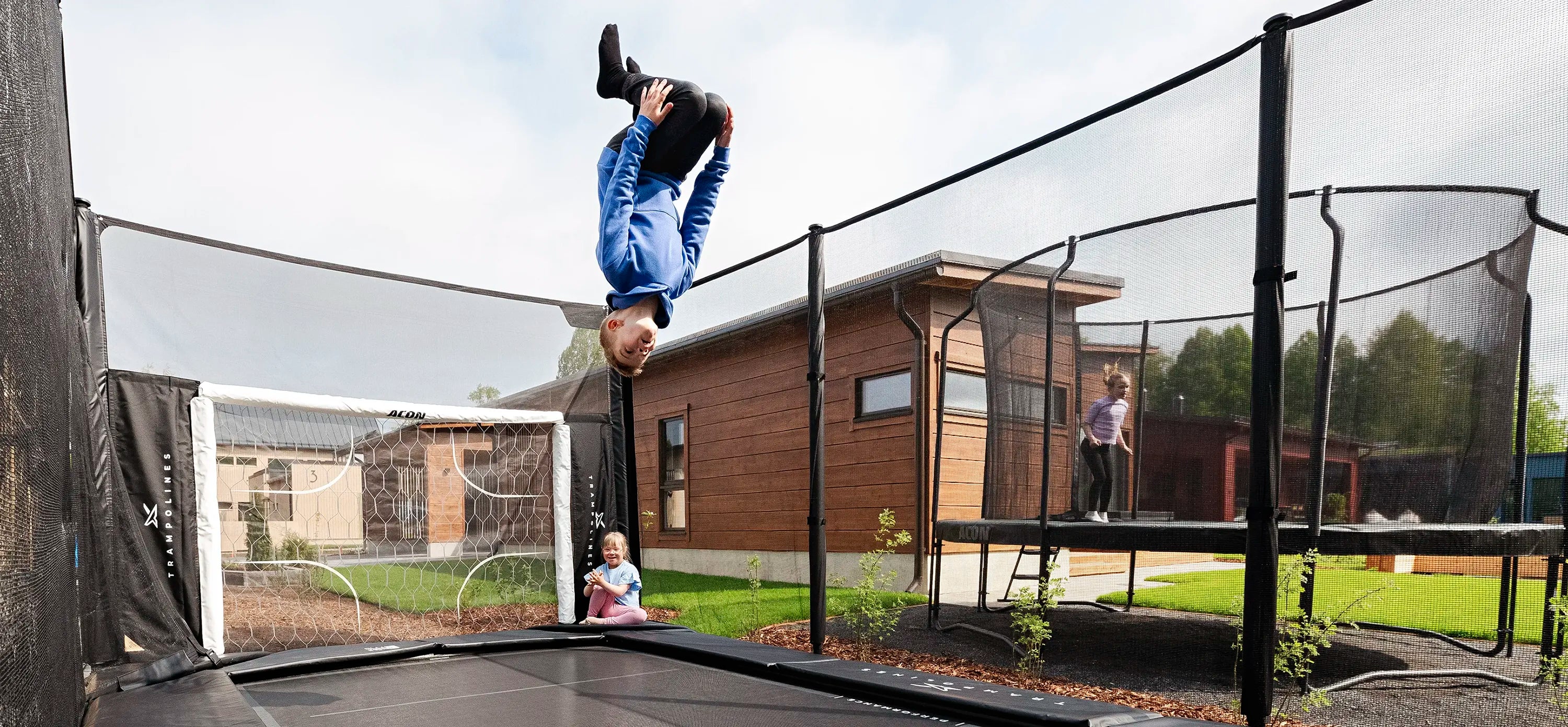Boy flips on Acon X trampoline, little girl claps. In the neighbor’s yard, a girl jumps on Acon Air trampoline.