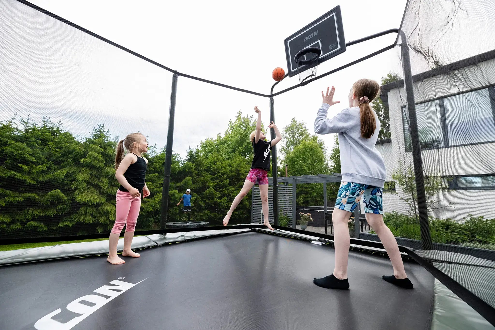 Kids playing basketball on a Acon trampoline with a basketball hoop in a backyard.