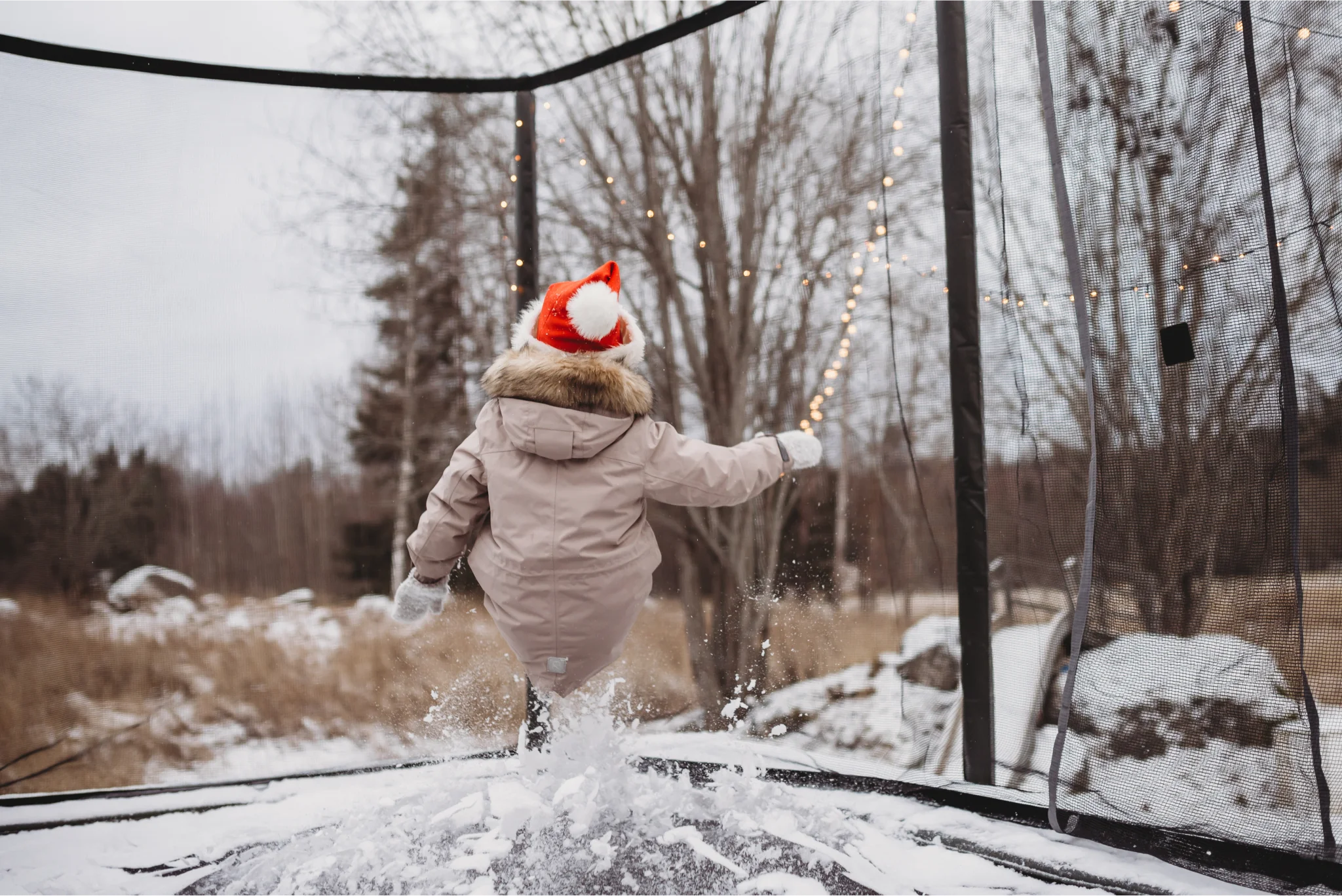 Child with a winter coat and a santa hat bouncing on a trampoline with snow.
