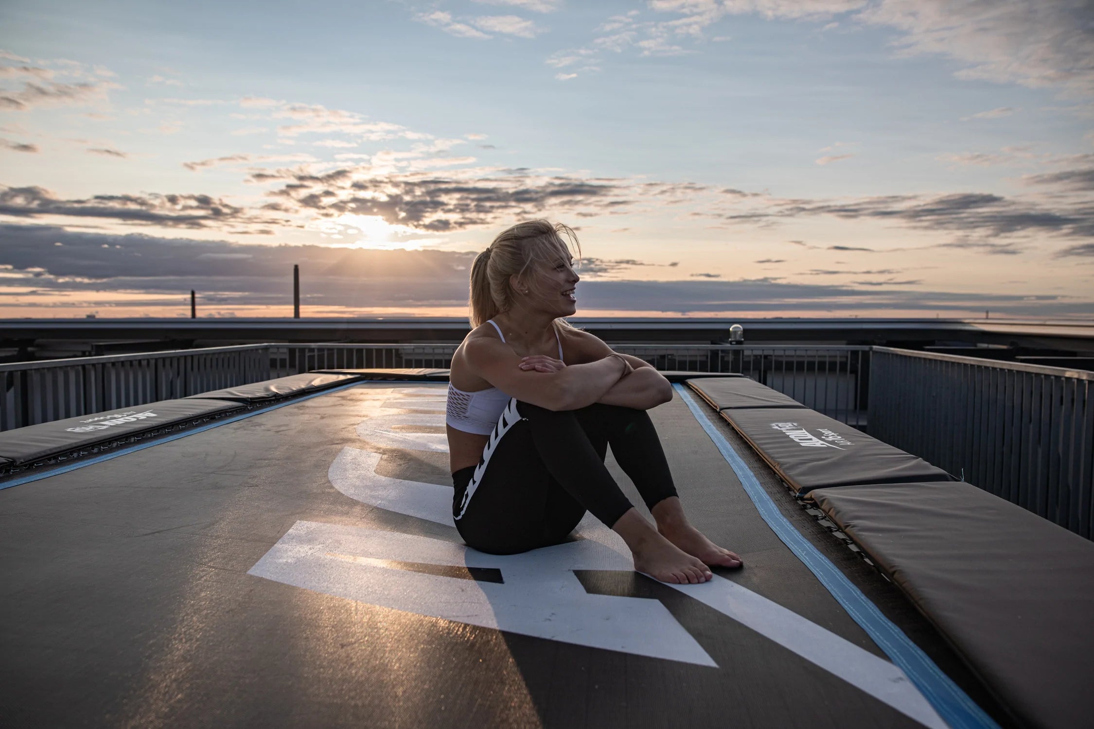 Cheerful woman sitting on an Acon 16 HD trampoline with a sunset as background