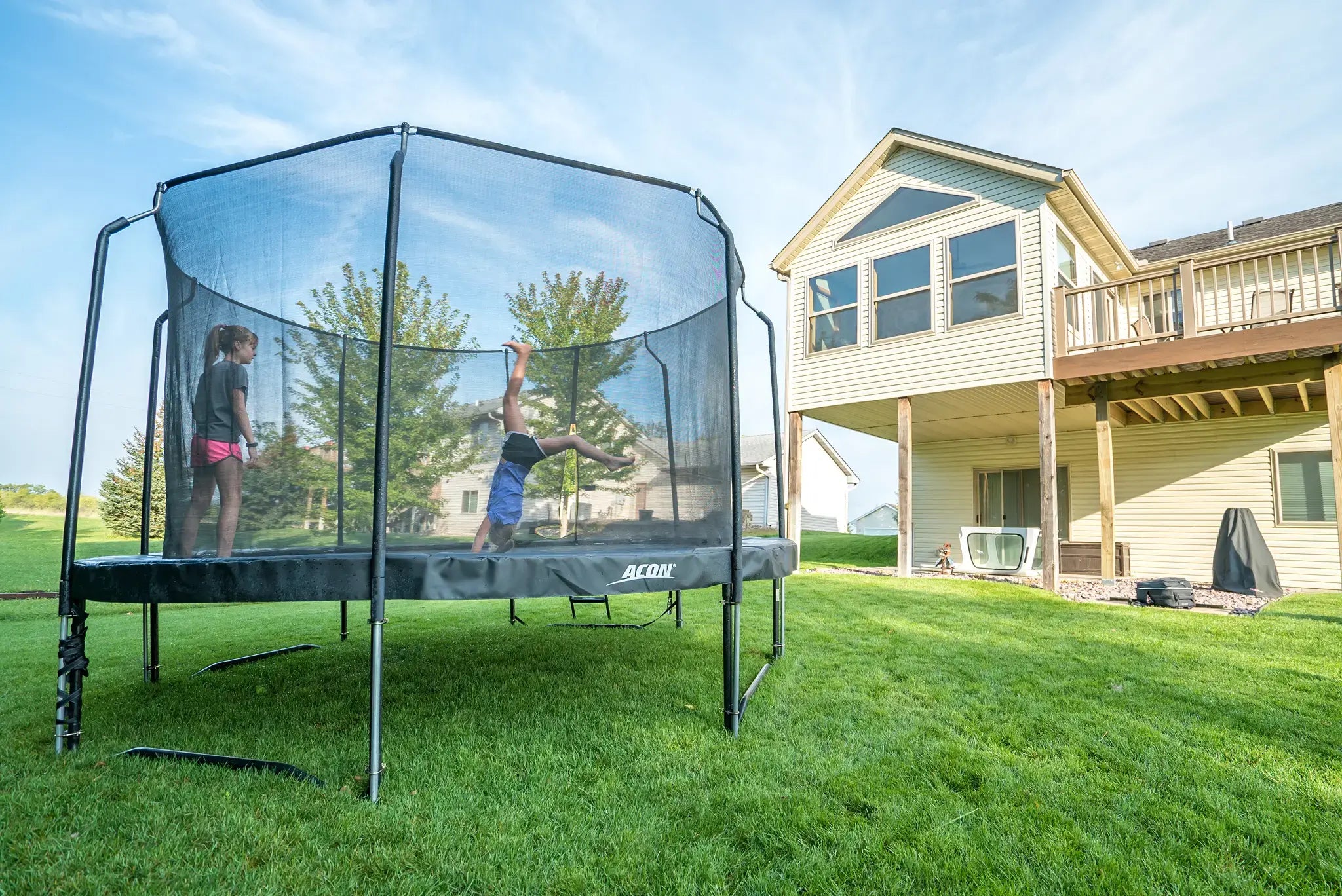 Two young girls on an Acon trampoline. Other one is doing a trick, while other watches from the side.