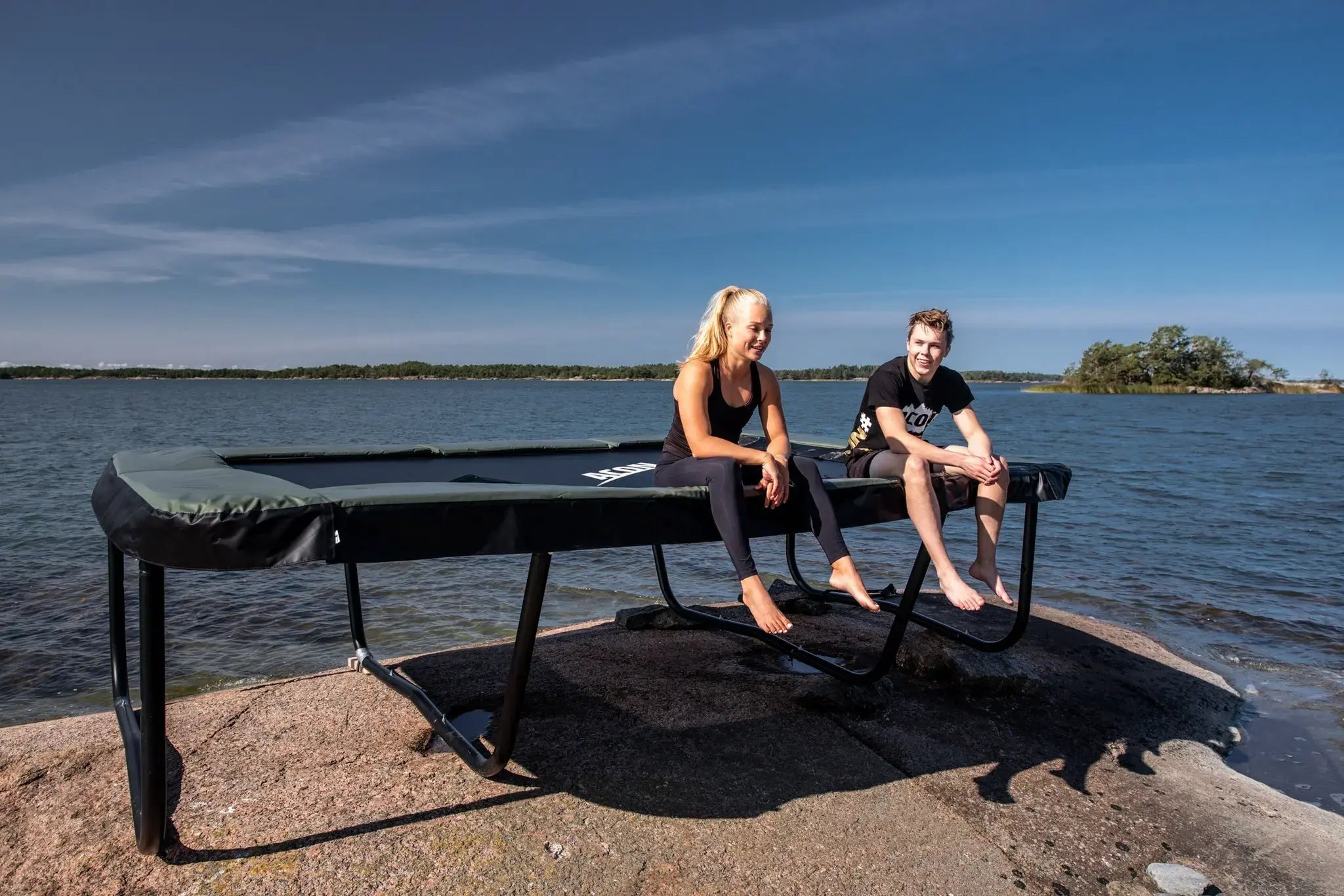 Two people sitting and relaxing on a rectangular Acon trampoline placed on a rocky shore by the sea under a clear blue sky.