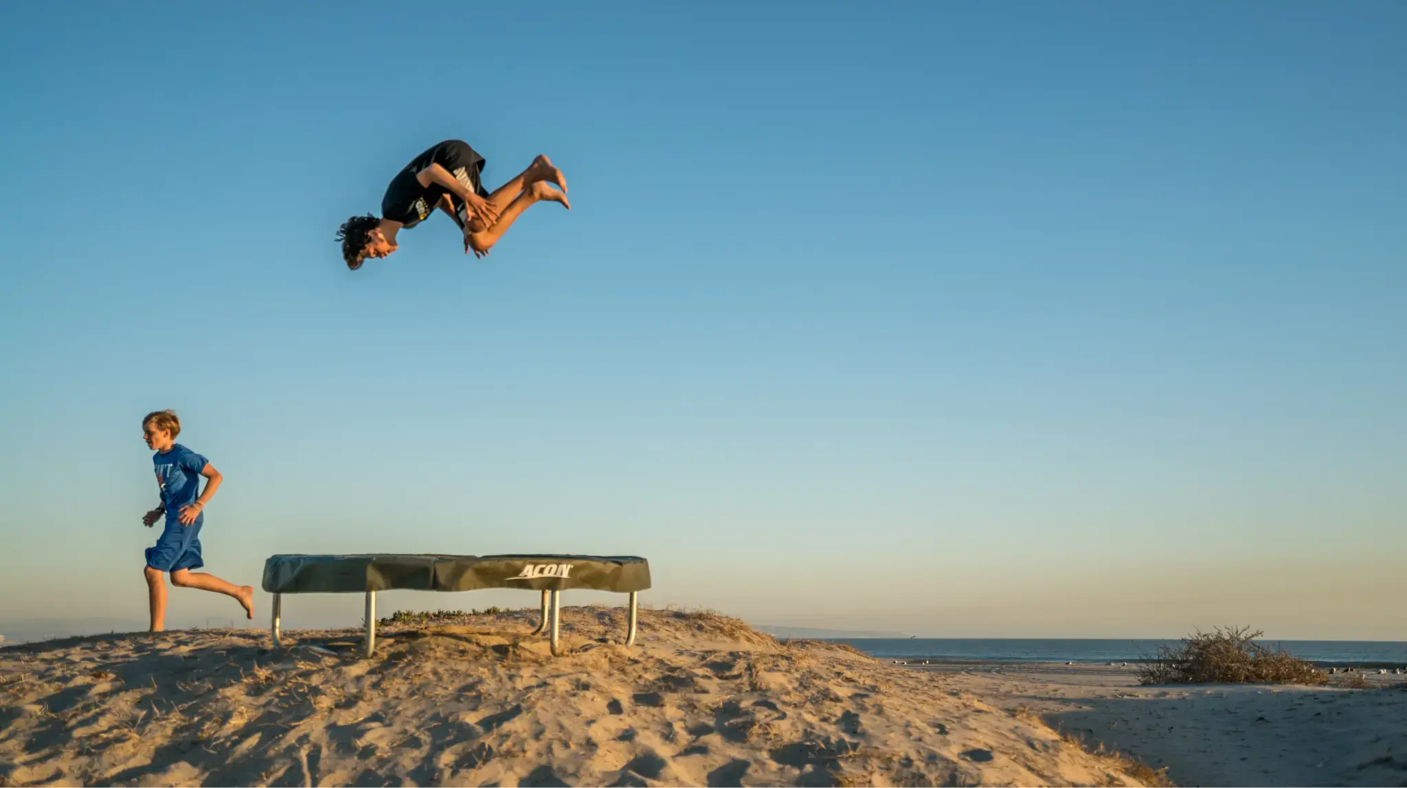 Two boys enjoying a trampoline on a sandy beach at sunset. One performs an acrobatic flip while the other runs nearby, with clear sky in the background.