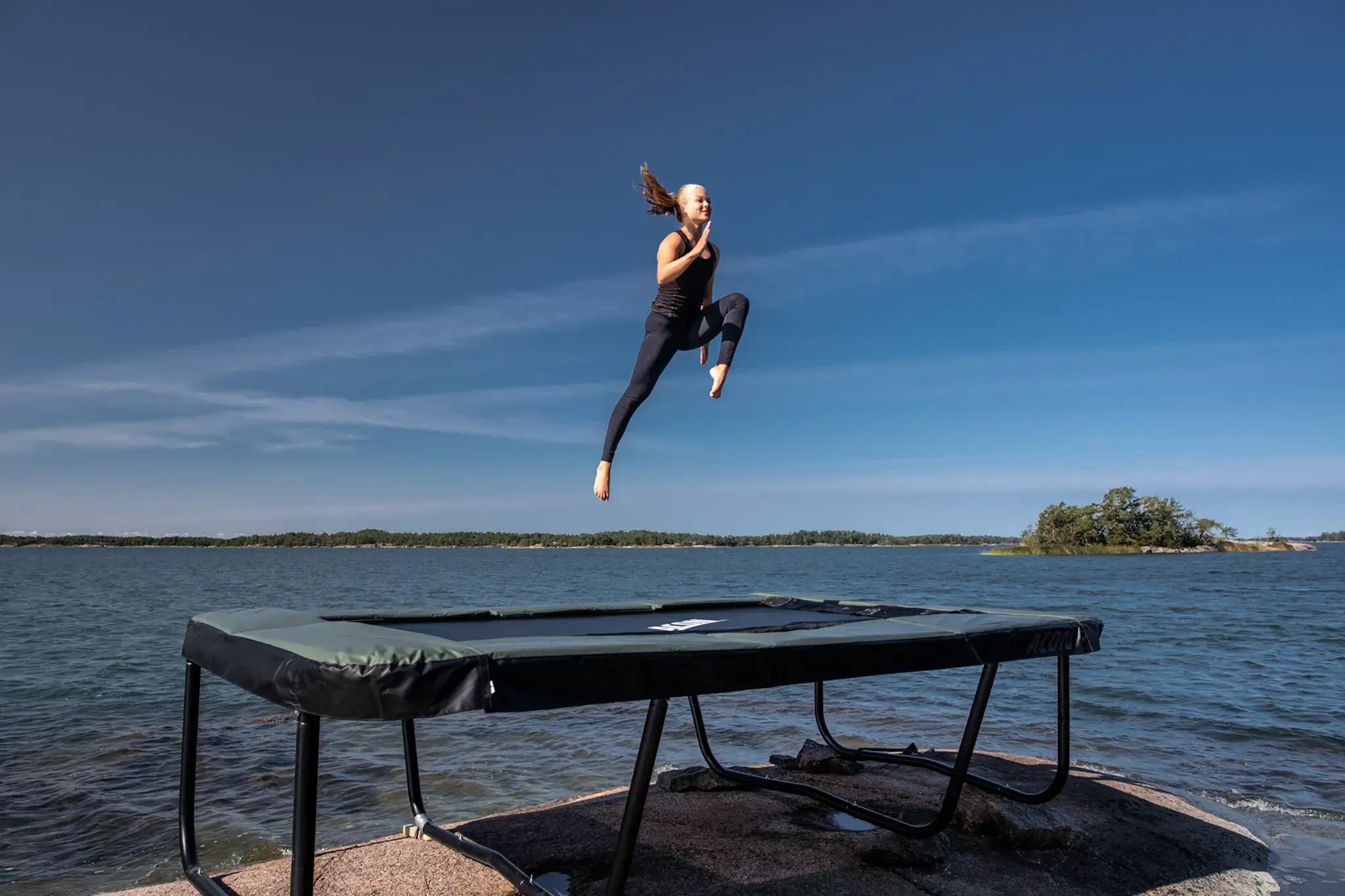 Person jumping high on a rectangular Acon trampoline set on a rocky shore by the sea under a clear blue sky.
