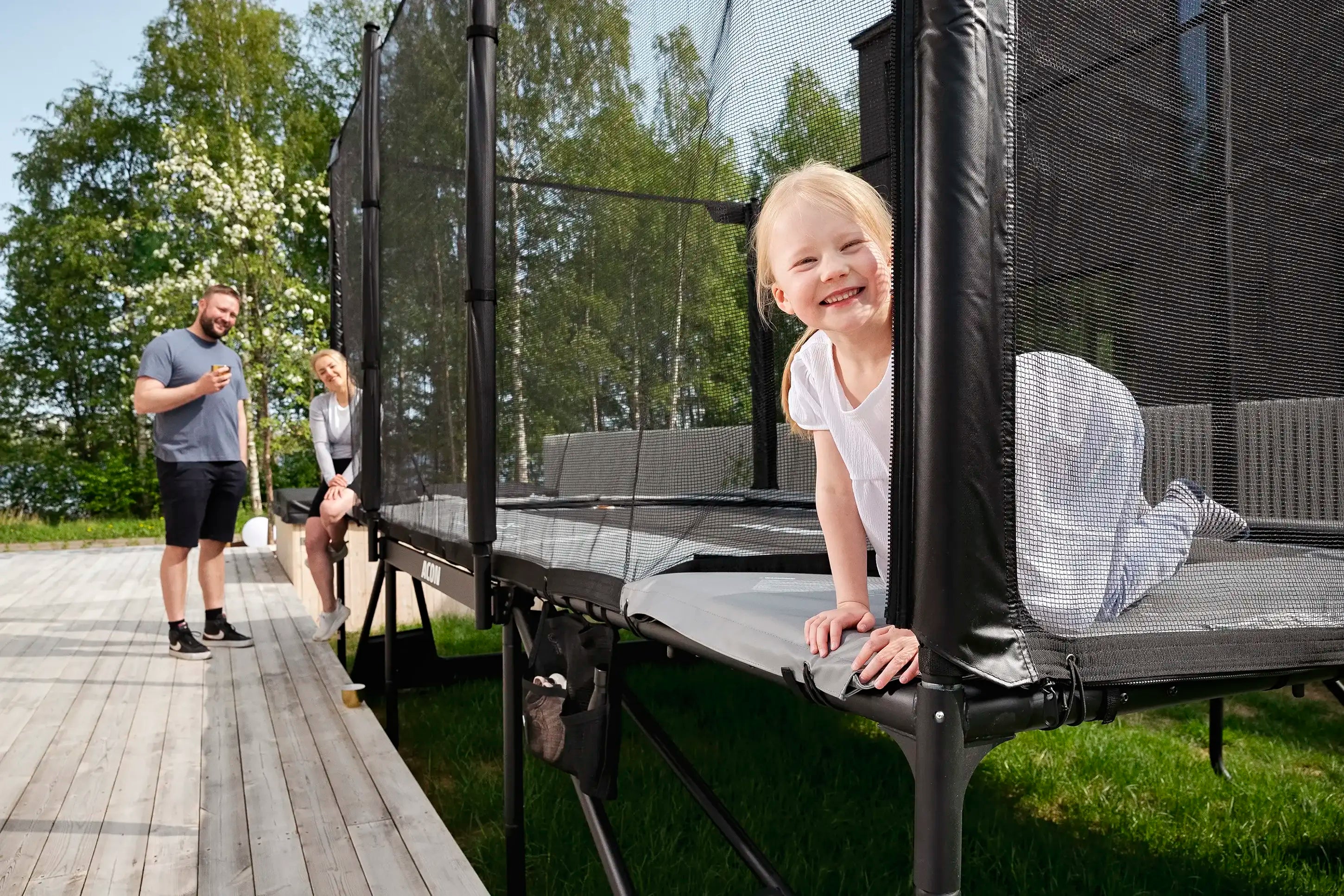 A little girl peeks through the Acon X trampoline netting, as her dad and mom stand just behind her, watching.