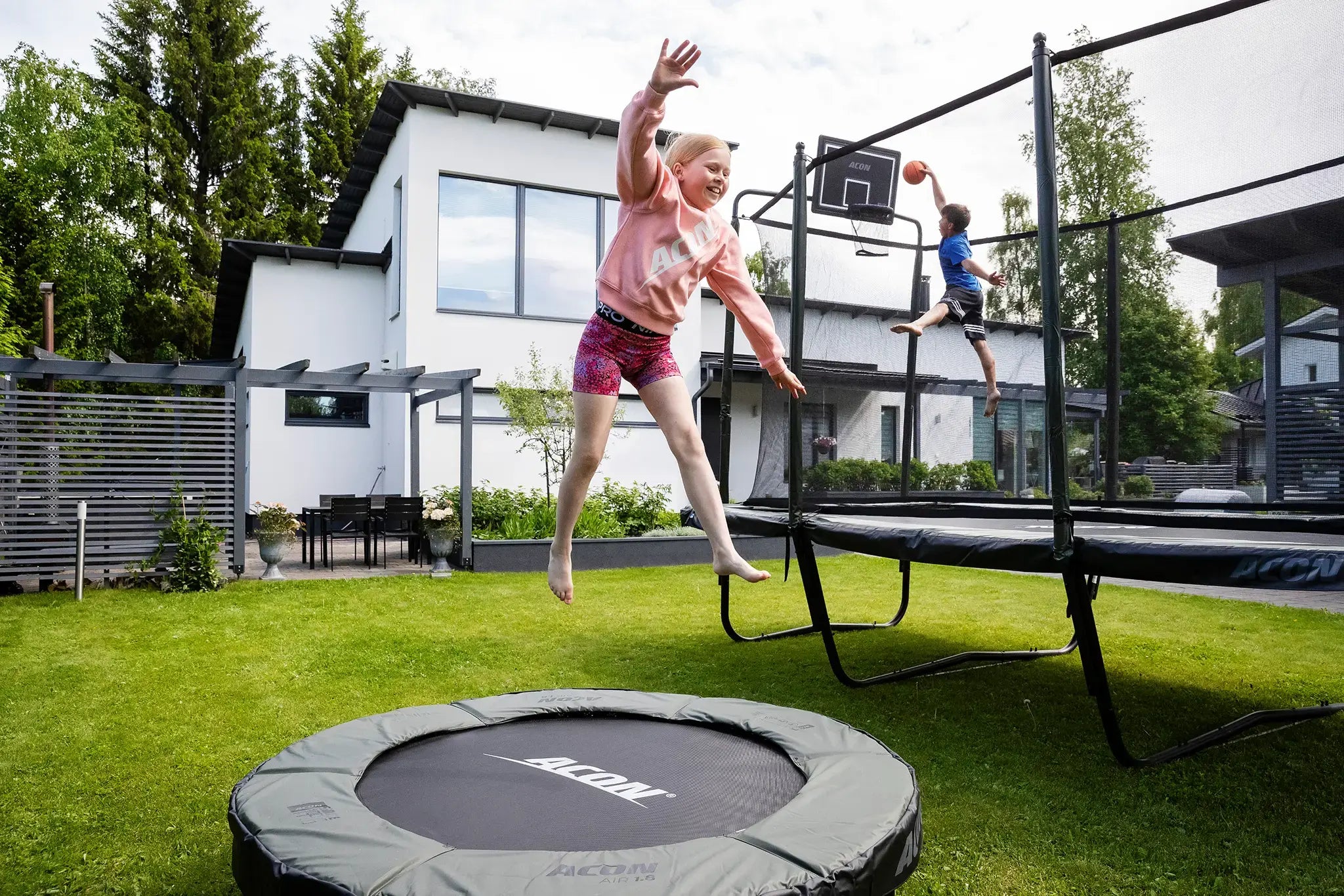 A boy is jumping and dunking a basketball on a bigger trampoline, while a girl is jumping on a smaller Acon trampoline in the foreground.