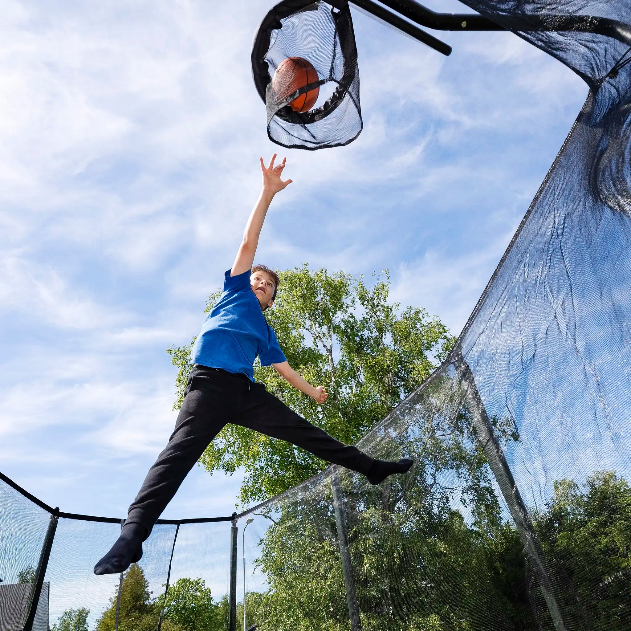 Boy jumping on a Acon trampoline and shooting a basketball into a hoop outdoors.