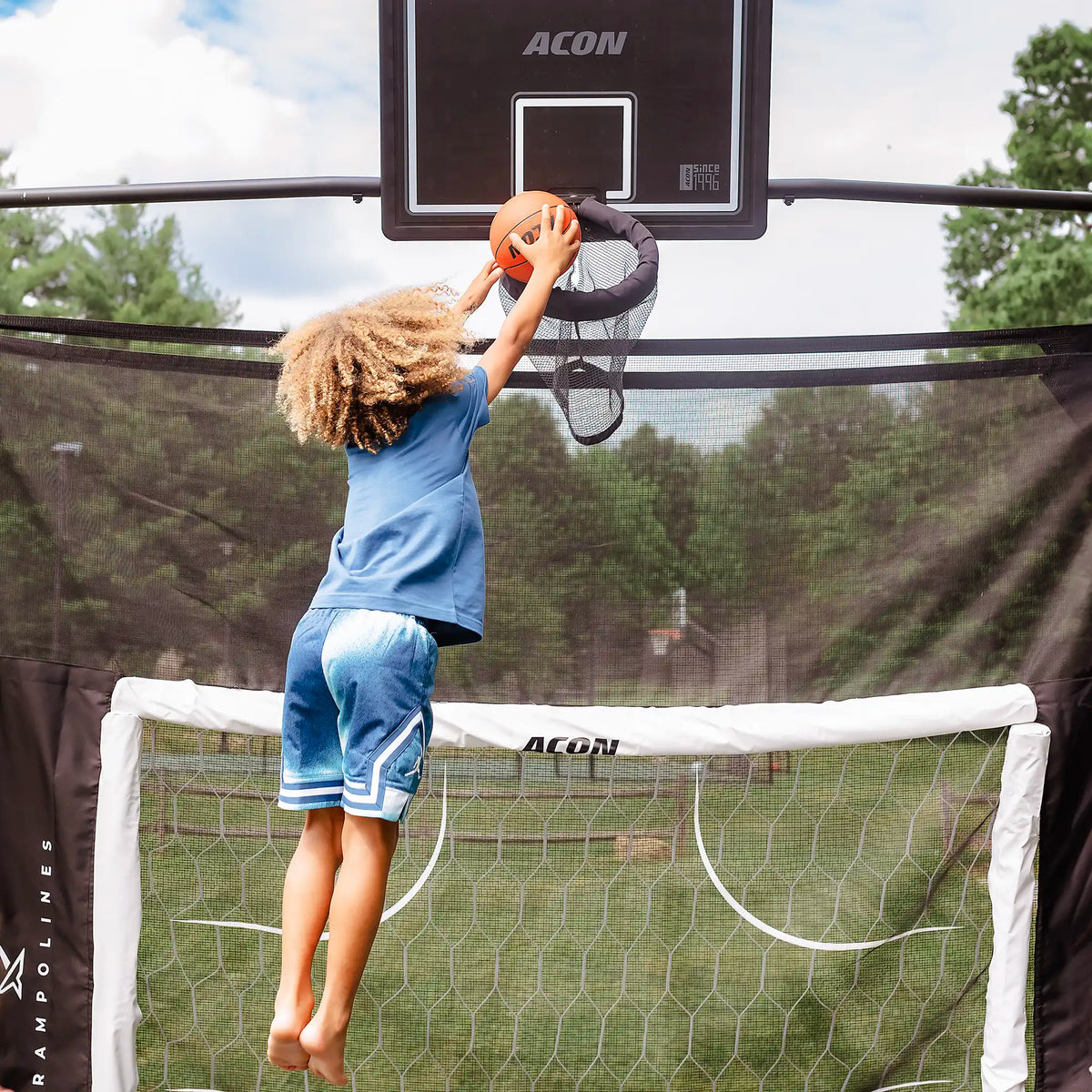 A child jumping on a trampoline while dunking a basketball into a hoop.
