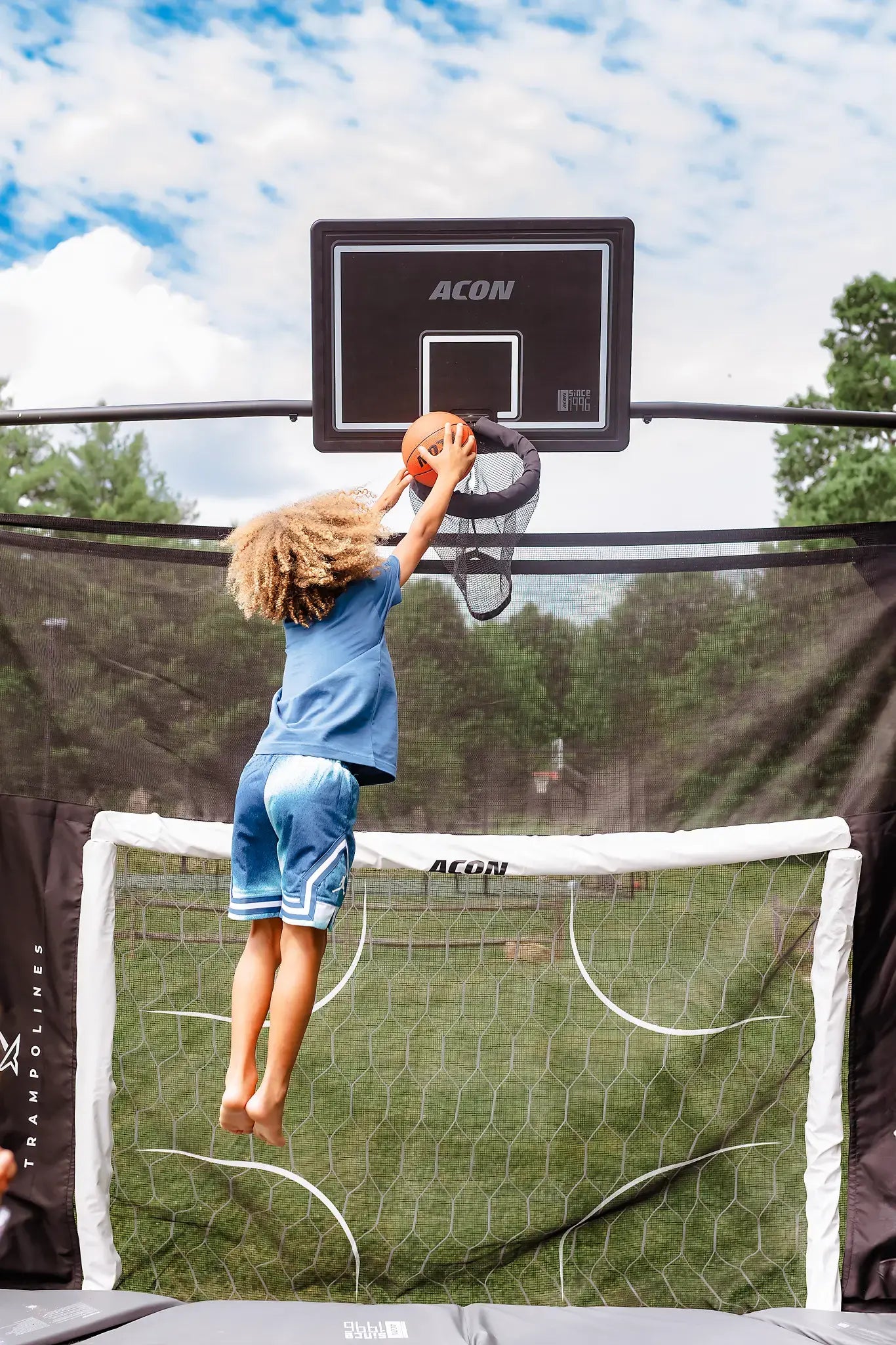 A child jumping on a trampoline while dunking a basketball into a hoop