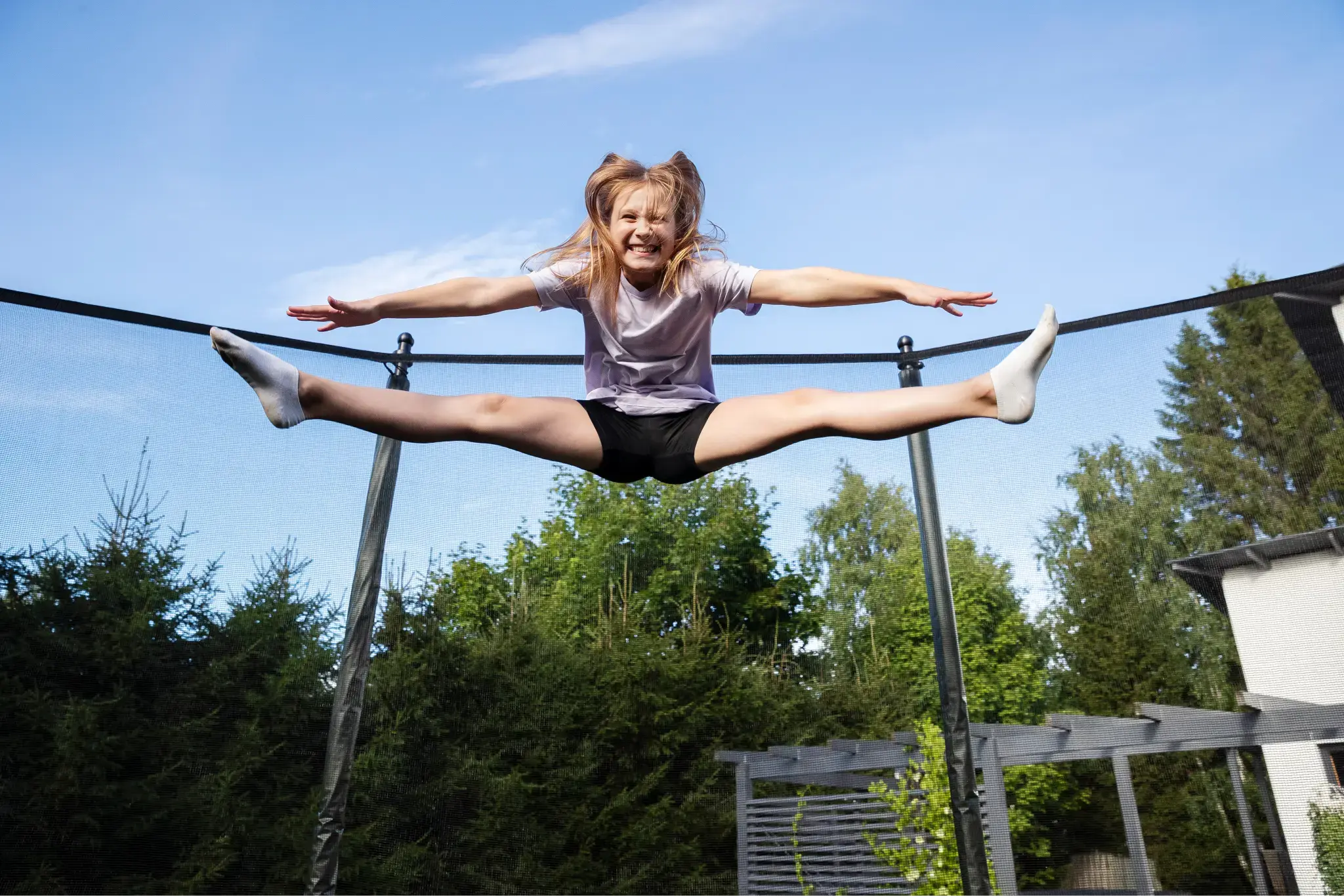 A smiling child performs a split jump on an Acon trampoline, jumping high against a clear blue sky.