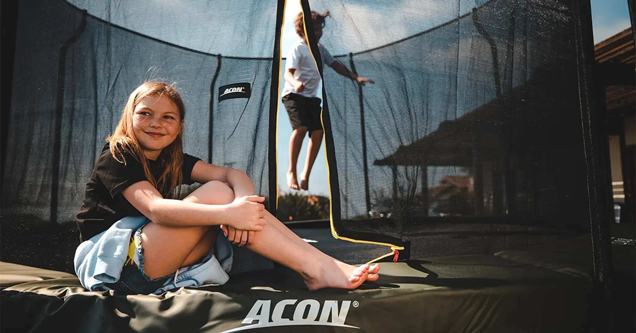 Girl sitting on the edge of a Acon trampoline while boy is jumping inside of it. 