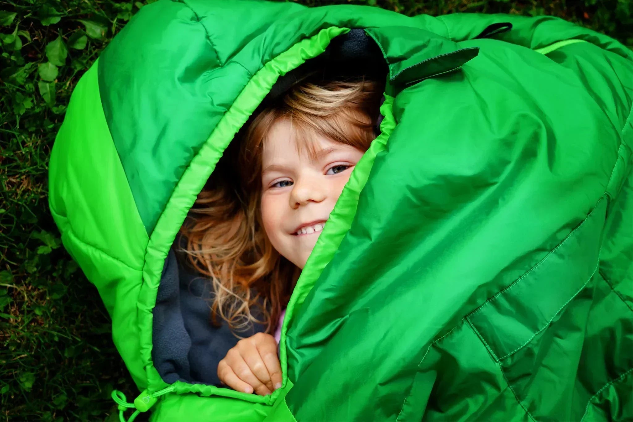 Kid peaking and smiling in a green sleeping bag while laying on the ground. 
