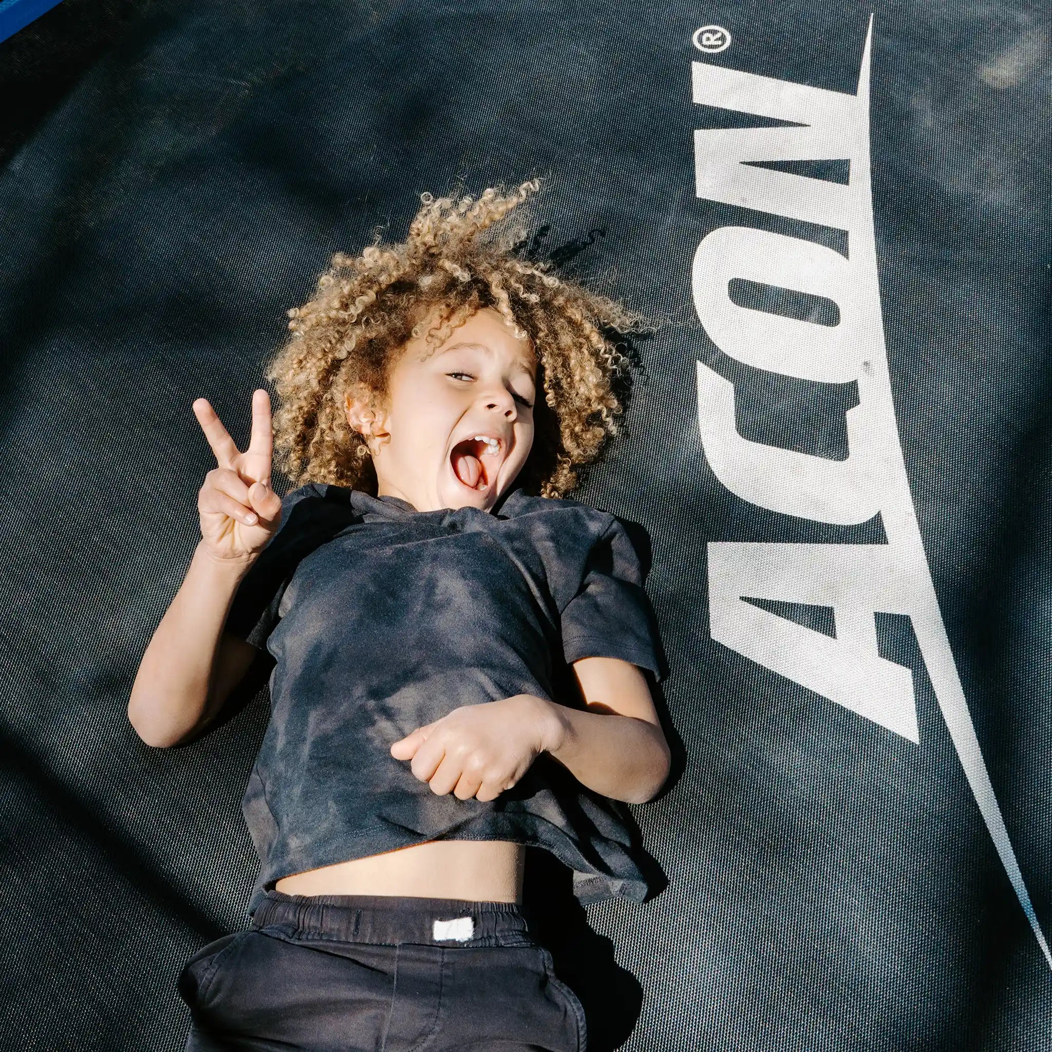 Young child laying on an Acon trampoline while smiling and showing the peace sign.