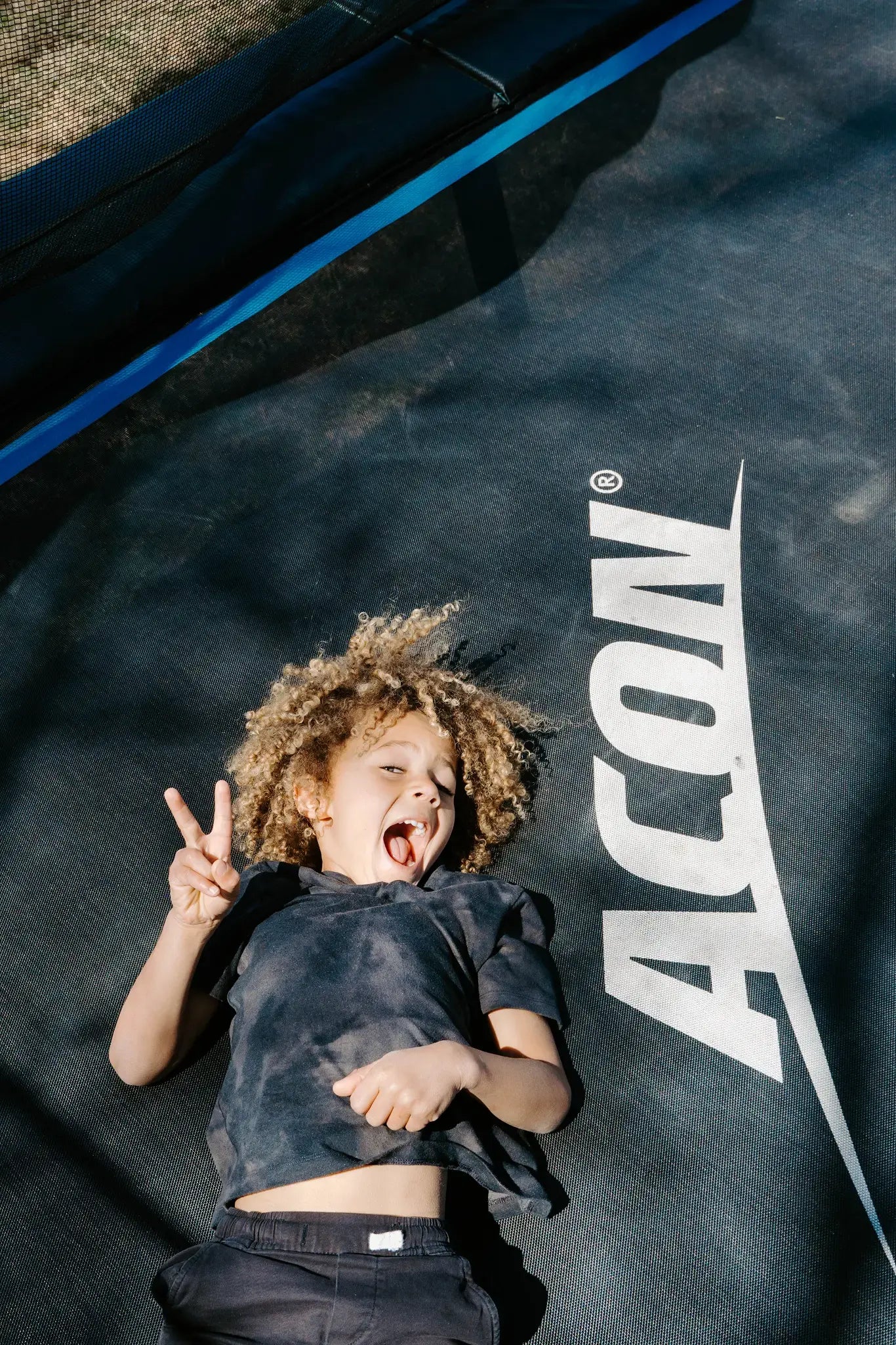 Young child laying on an Acon trampoline while smiling and showing the peace sign. 