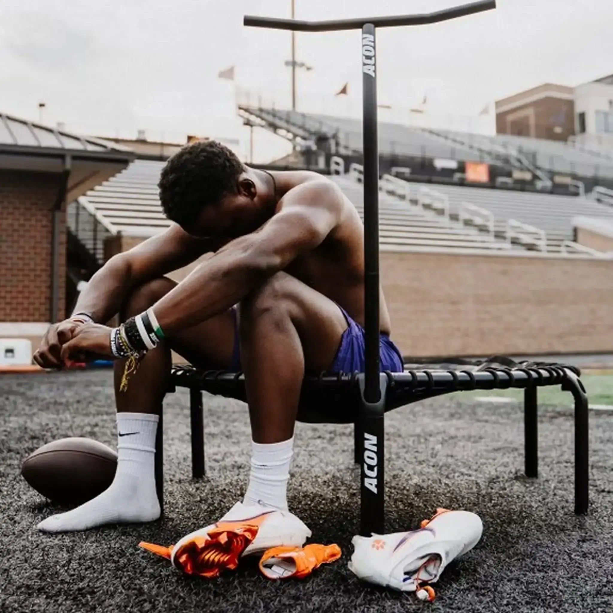An athletic man sits on an ACON rebounder trampoline resting. His football cleats, gloves, and a football lie on the ground beside him.
