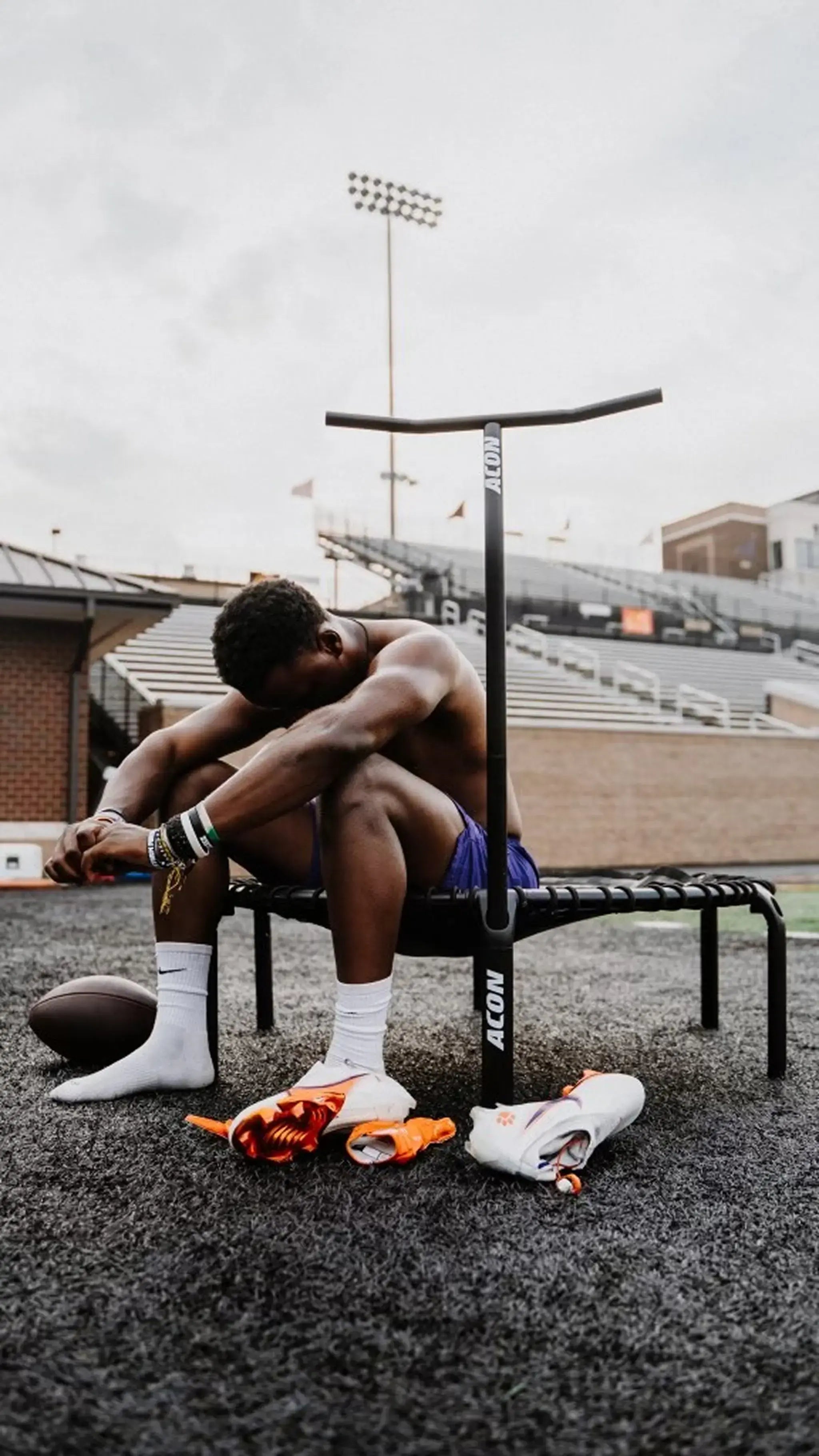 An athletic man sits on an ACON rebounder trampoline resting. His football cleats, gloves, and a football lie on the ground beside him.