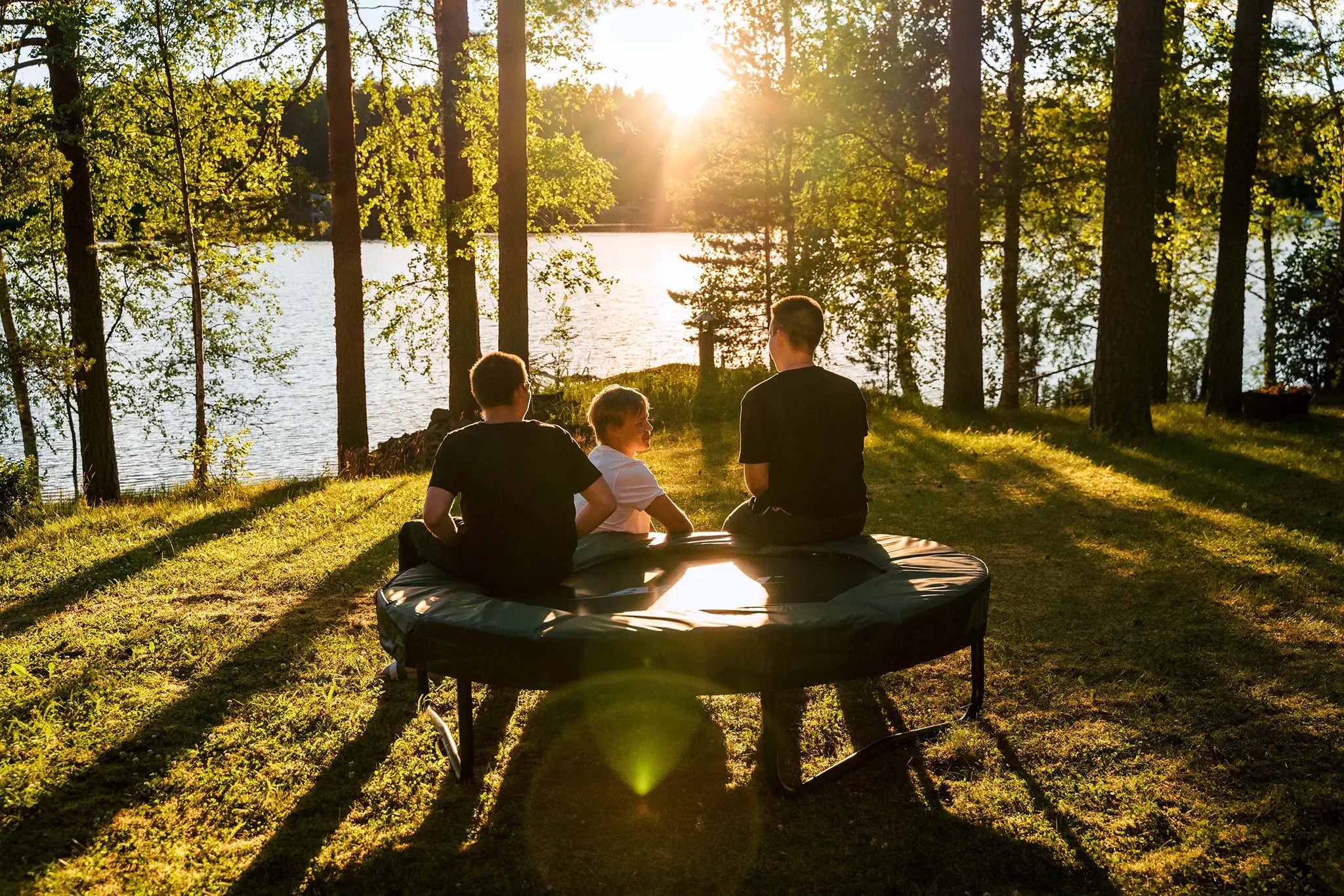 Three boys sitting on an Acon trampoline by a lakeside at sunset, enjoying a peaceful summer evening.