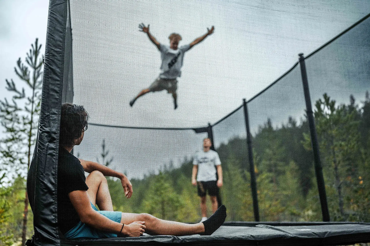 Person jumping high on a trampoline while two others sit and stand nearby inside the safety enclosure.
