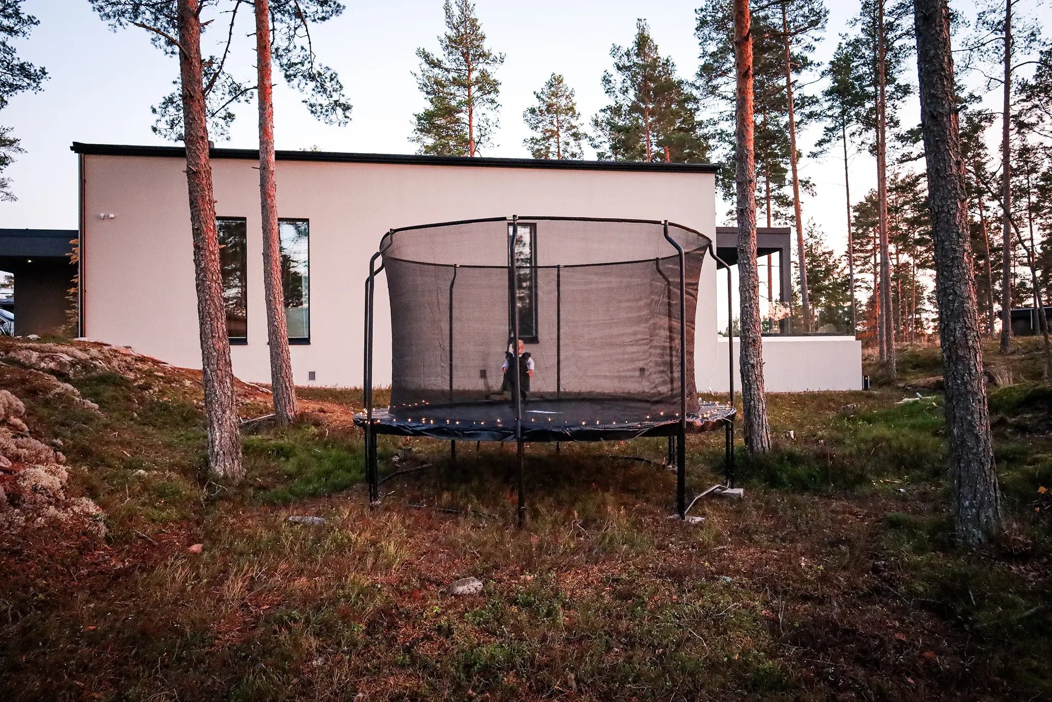 Child standing inside an Acon trampoline with a safety net, set up in a forested yard at sunset with string lights decorating the frame.