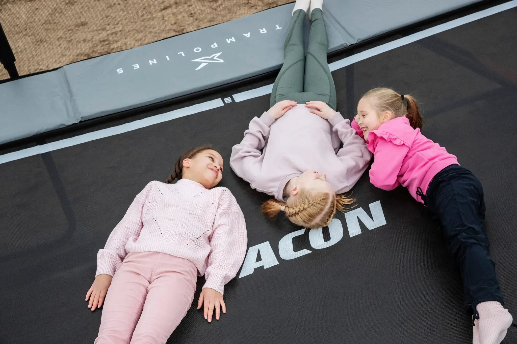 Three young girls lying on an Acon trampoline, smiling and chatting.