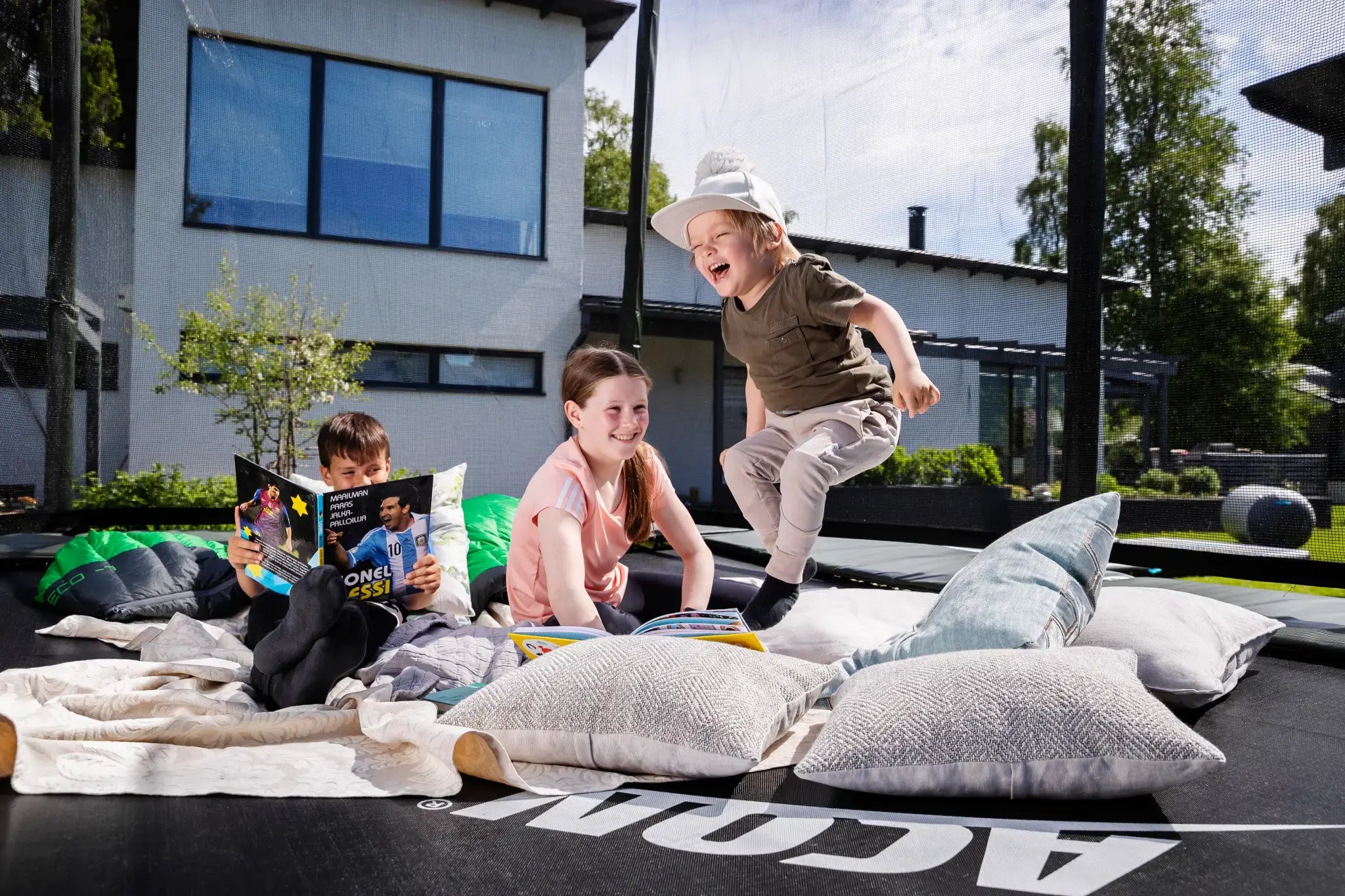 Three children enjoying a trampoline setup with blankets and pillows. One reads a book, another smiles, and a younger child joyfully jumps, with a modern house in the background.