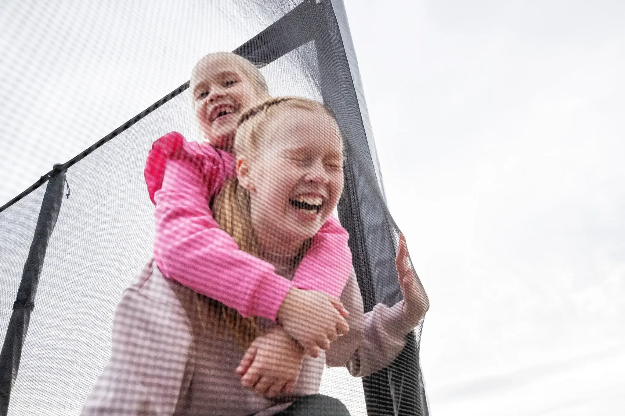 Two laughing girls playing on a Acon trampoline, seen through the safety net, with bright pink sweaters adding a playful vibe.