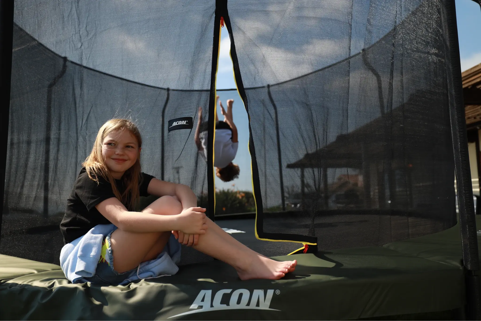 A smiling girl sits on the edge of a Acon trampoline, with another child doing a flip inside the trampoline in the background, on a sunny day.