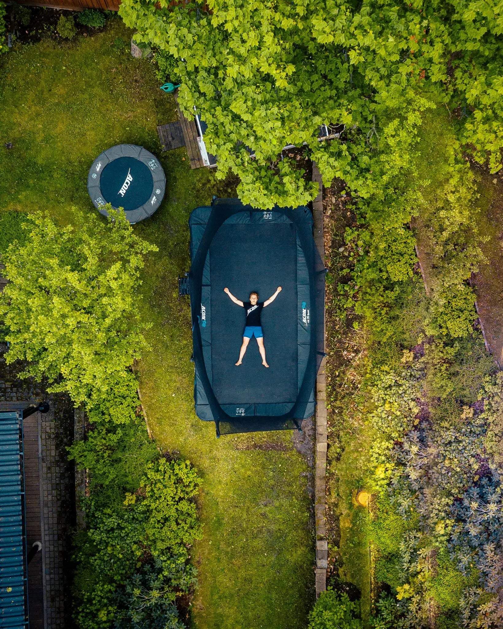 A boy lying on Acon 16 Sport HD trampoline in a backyard, seen above. 