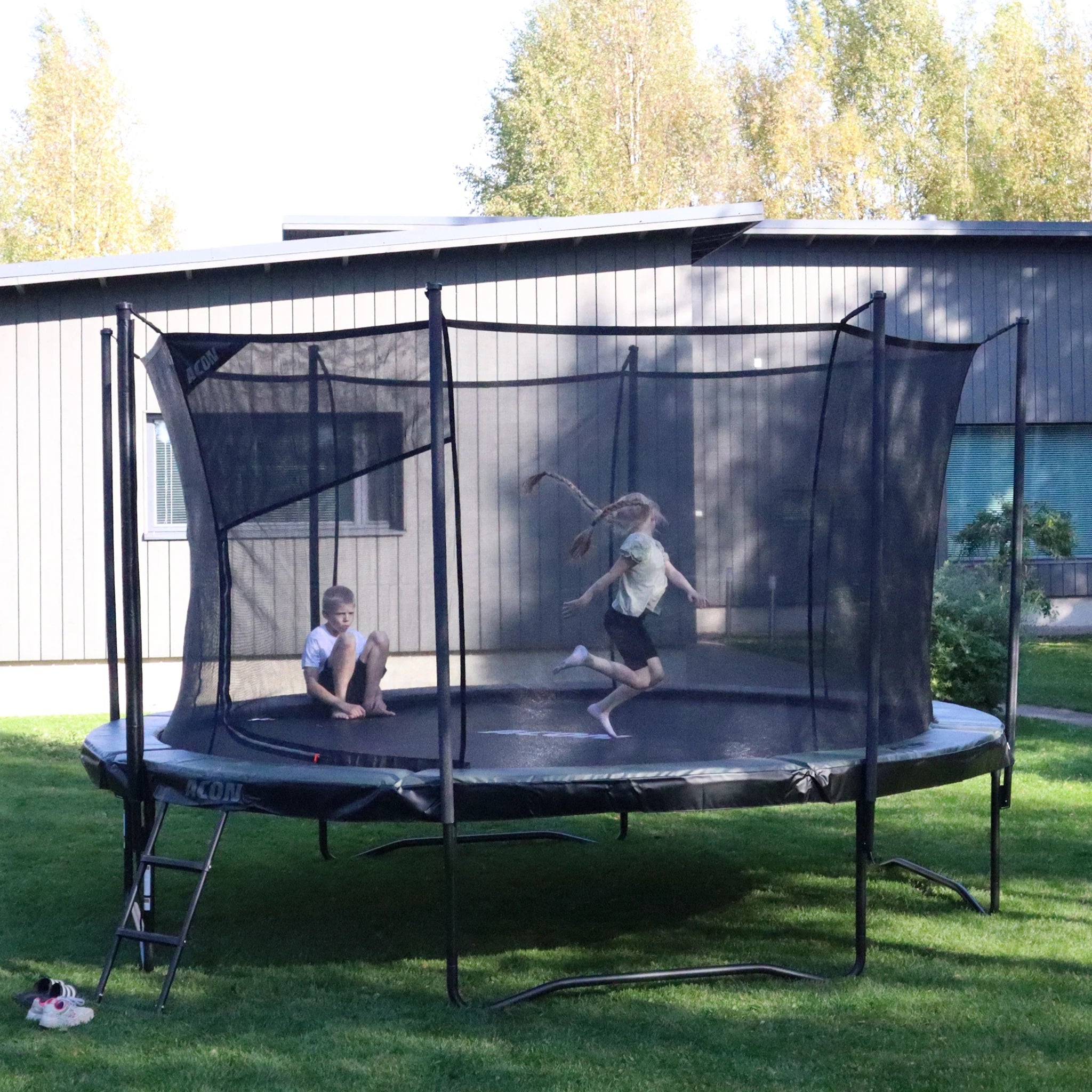 Acon Air trampoline in a detached house backyard. A boy sits on the trampoline while a girl is jumping.