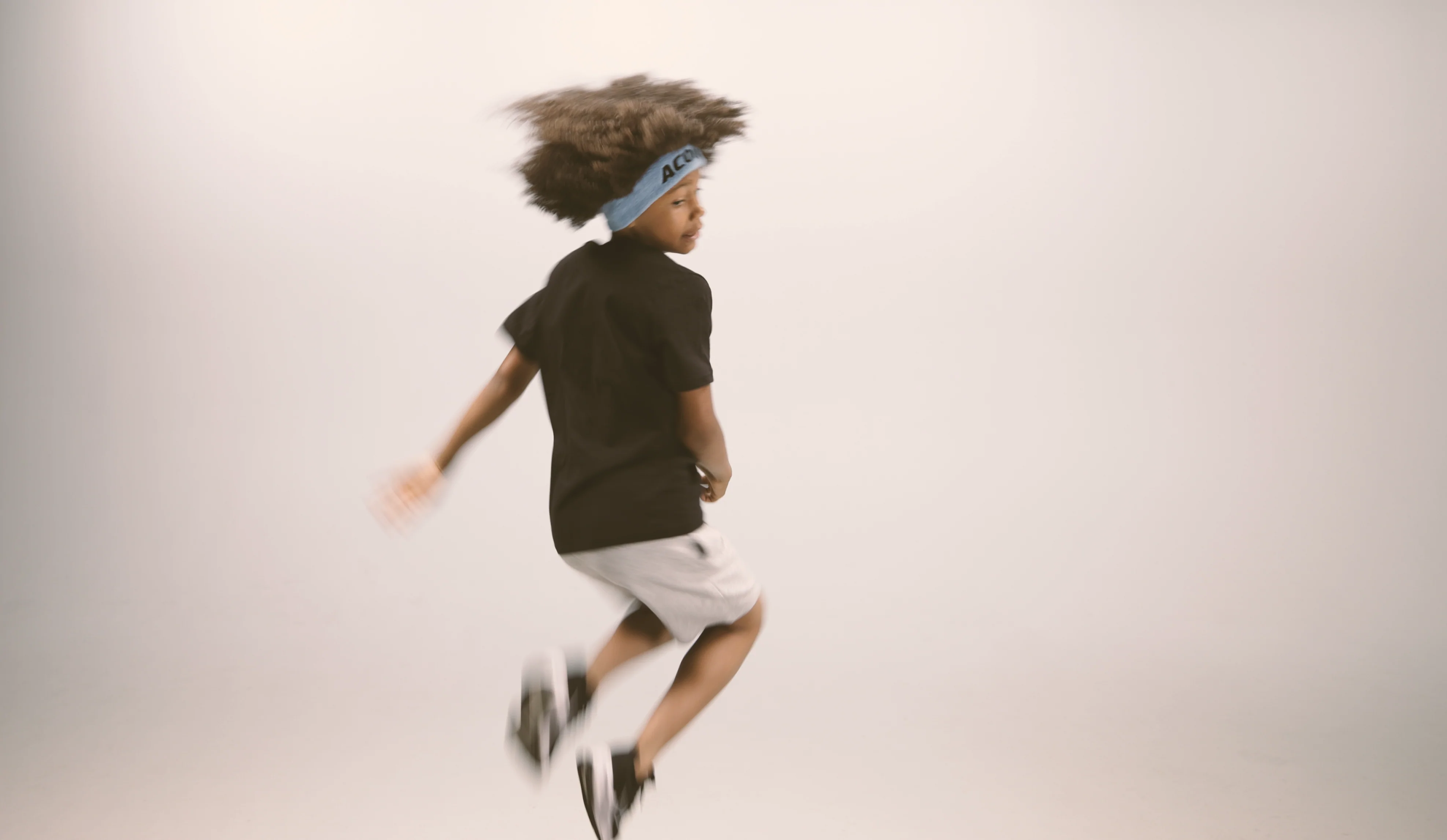 A jumping kid wearing an Acon headband, black t-shirt, white shorts and sneakers. Image is taken against beige background 