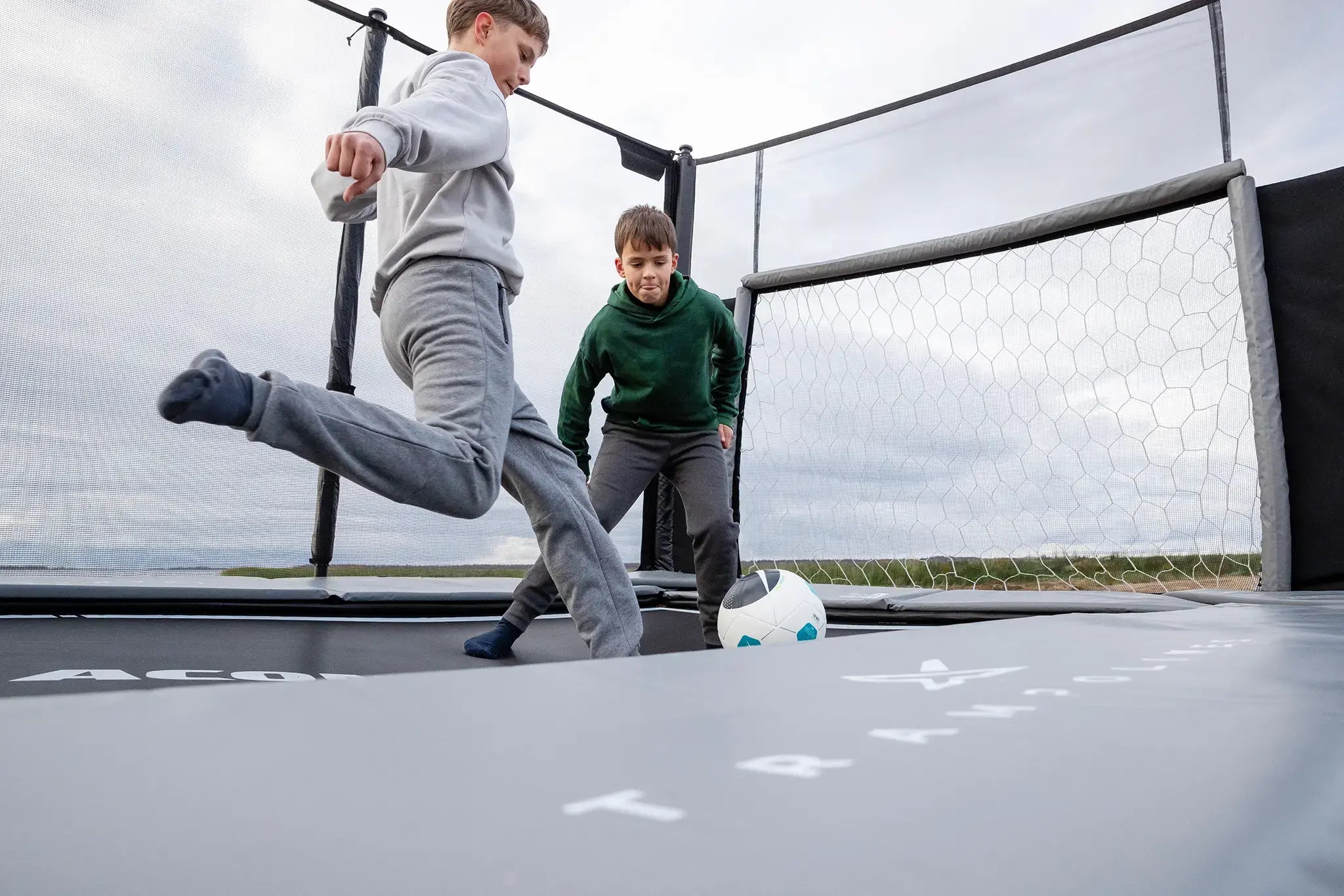 Two boys playing soccer on a Acon trampoline with a goal net.