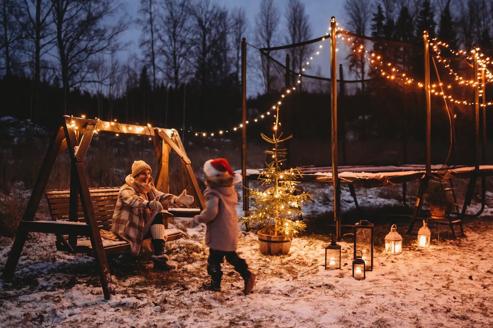 Winter backyard  with an Acon trampoline decorated with string lights, an adult sitting on a swing, and a child running towards her. 