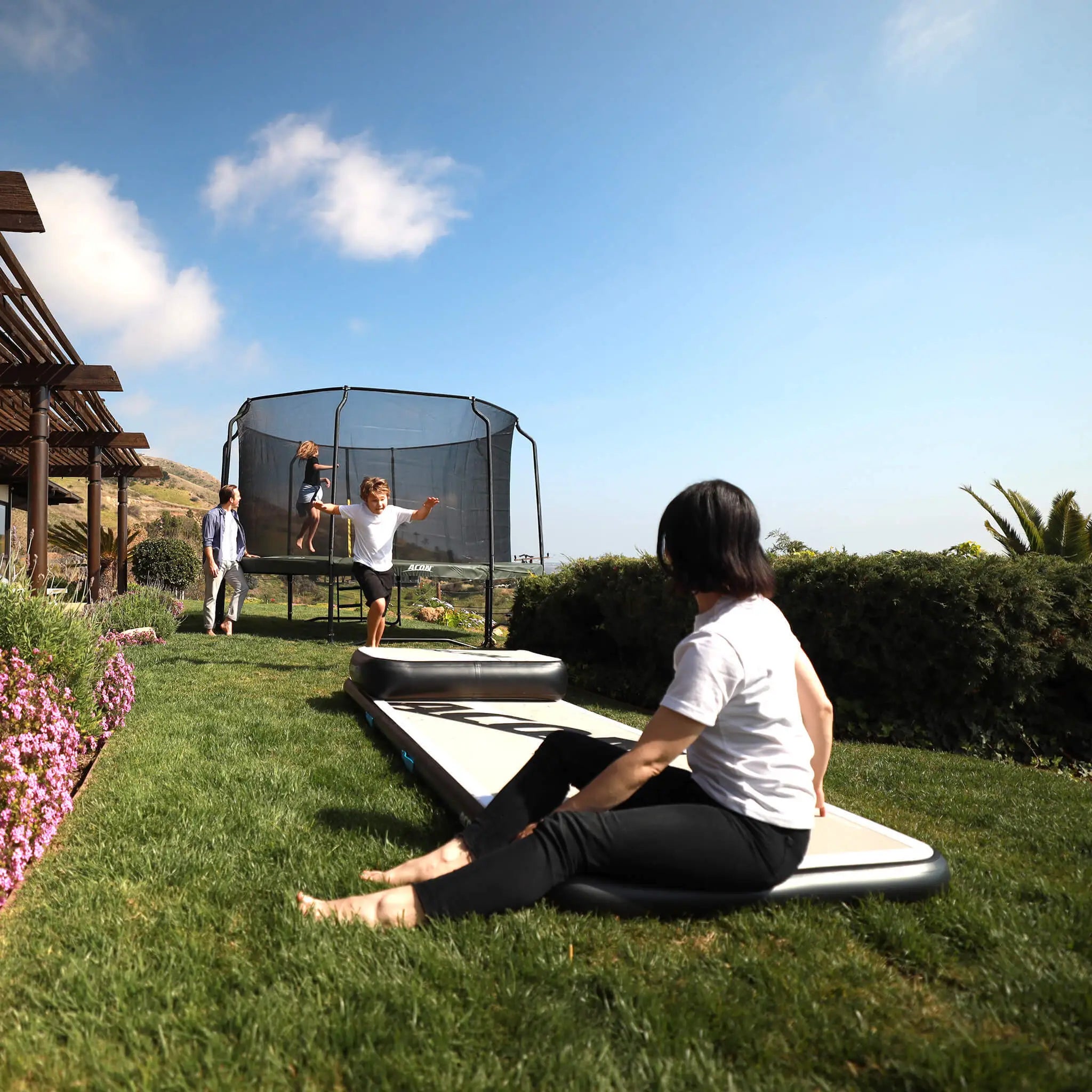 Family playing in the garden, girl jumping on a trampoline, boy running towards an airtrack. 