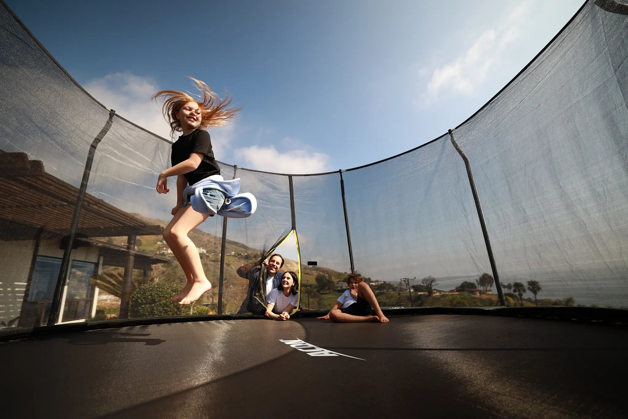 Girl jumping on an Acon trampoline with a safety net while family members watch.