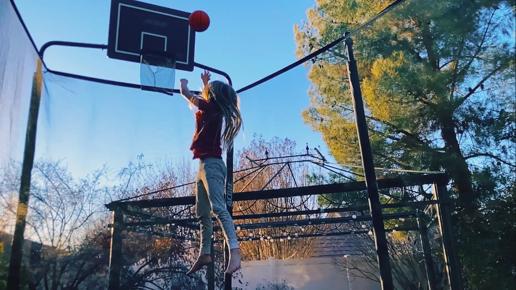 Young girl jumping on a trampoline while shooting a basketball into a acon basketball hoop.