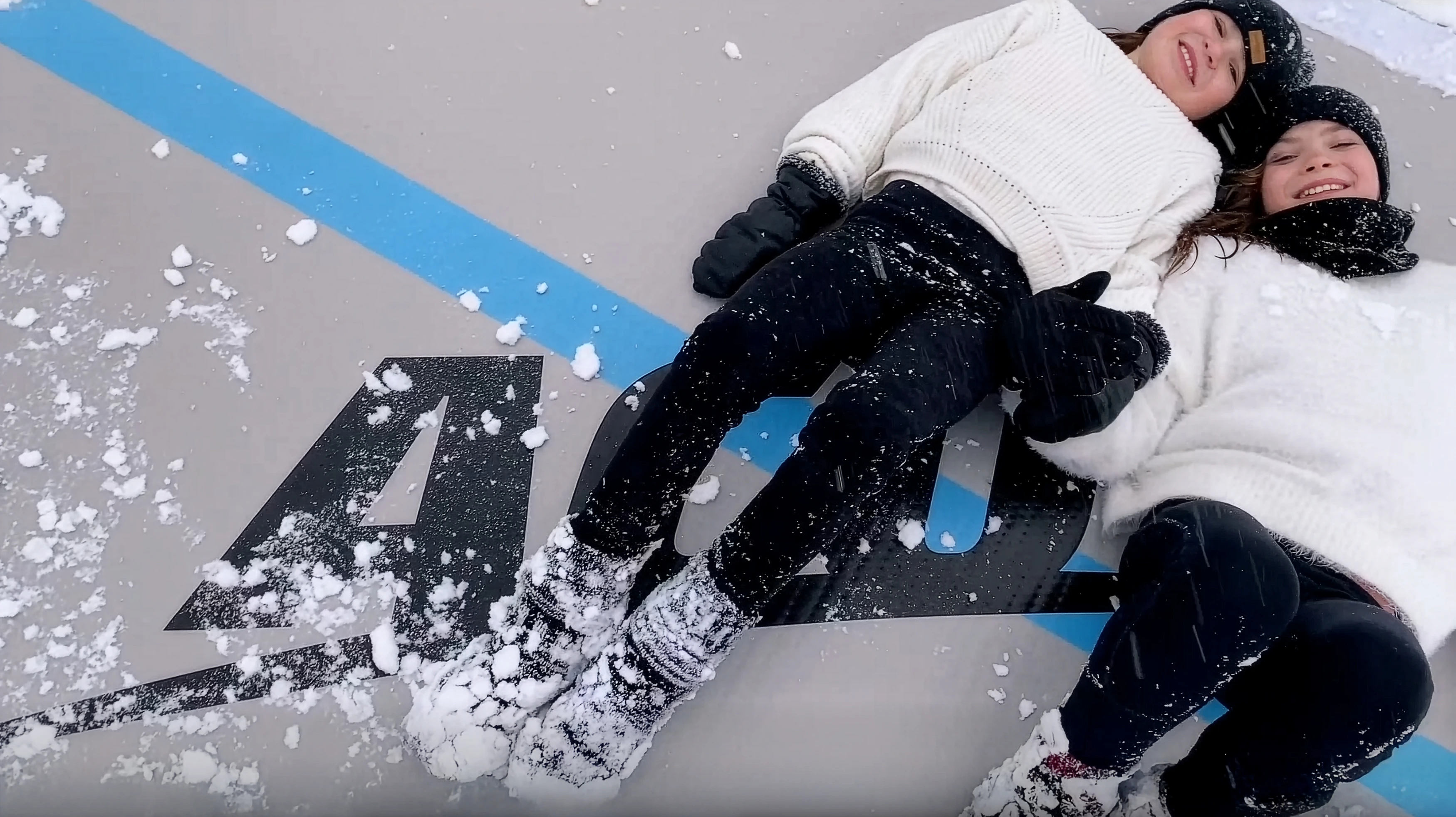 Two smiling sisters lying side by side on an Acon airtrack with a light dusting of snow on it.