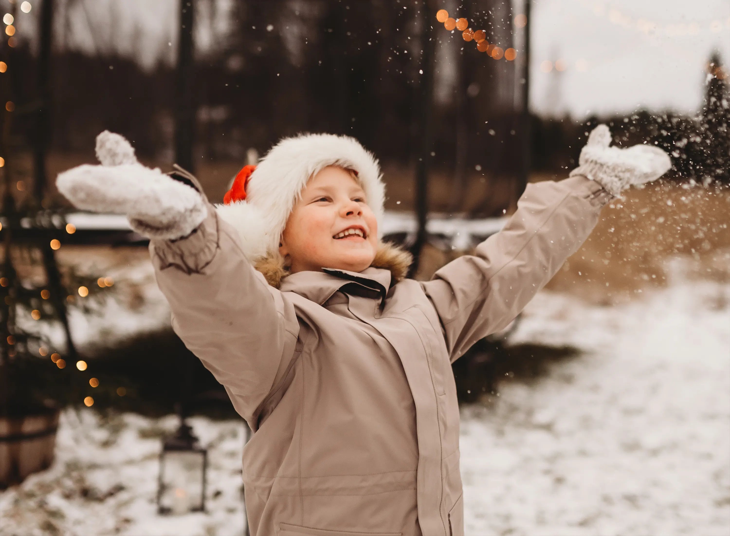 Child in snowy outdoor - in front of the 16 HD trampoline - setting with arms outstretched, wearing a beige coat and Santa hat.