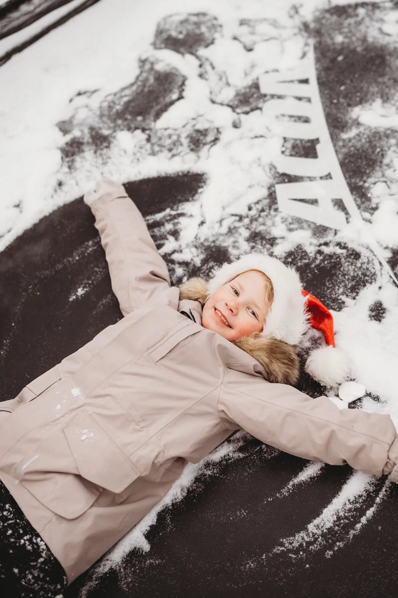 Child making a snow angel on a snowy Acon trampoline wearing a Santa hat and winter coat.