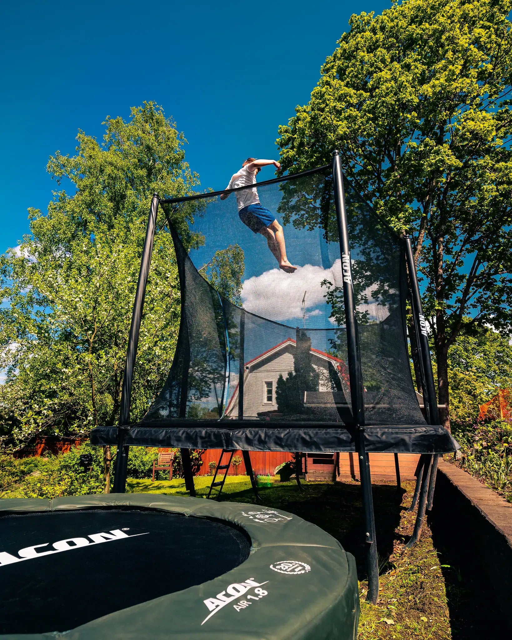 Person jumping high on an Acon  trampoline in a sunny backyard surrounded by trees.