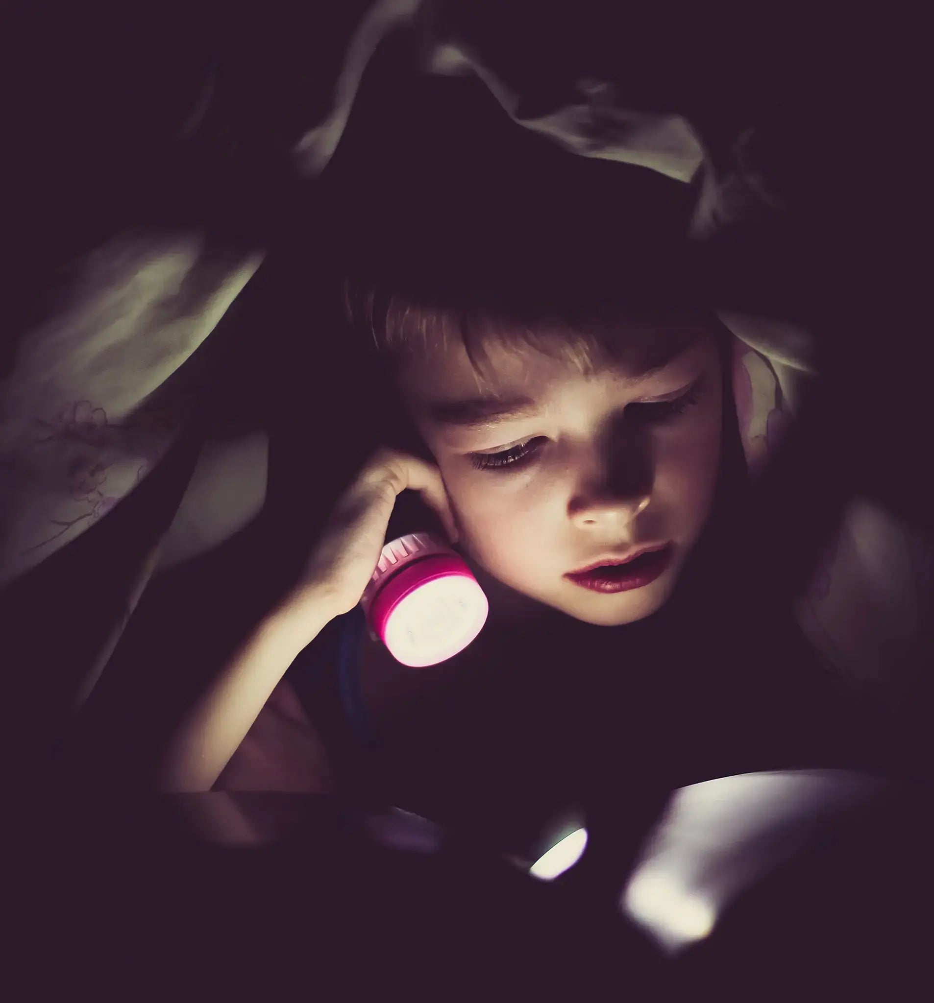 Kid reading with a flashlight in the dark. 