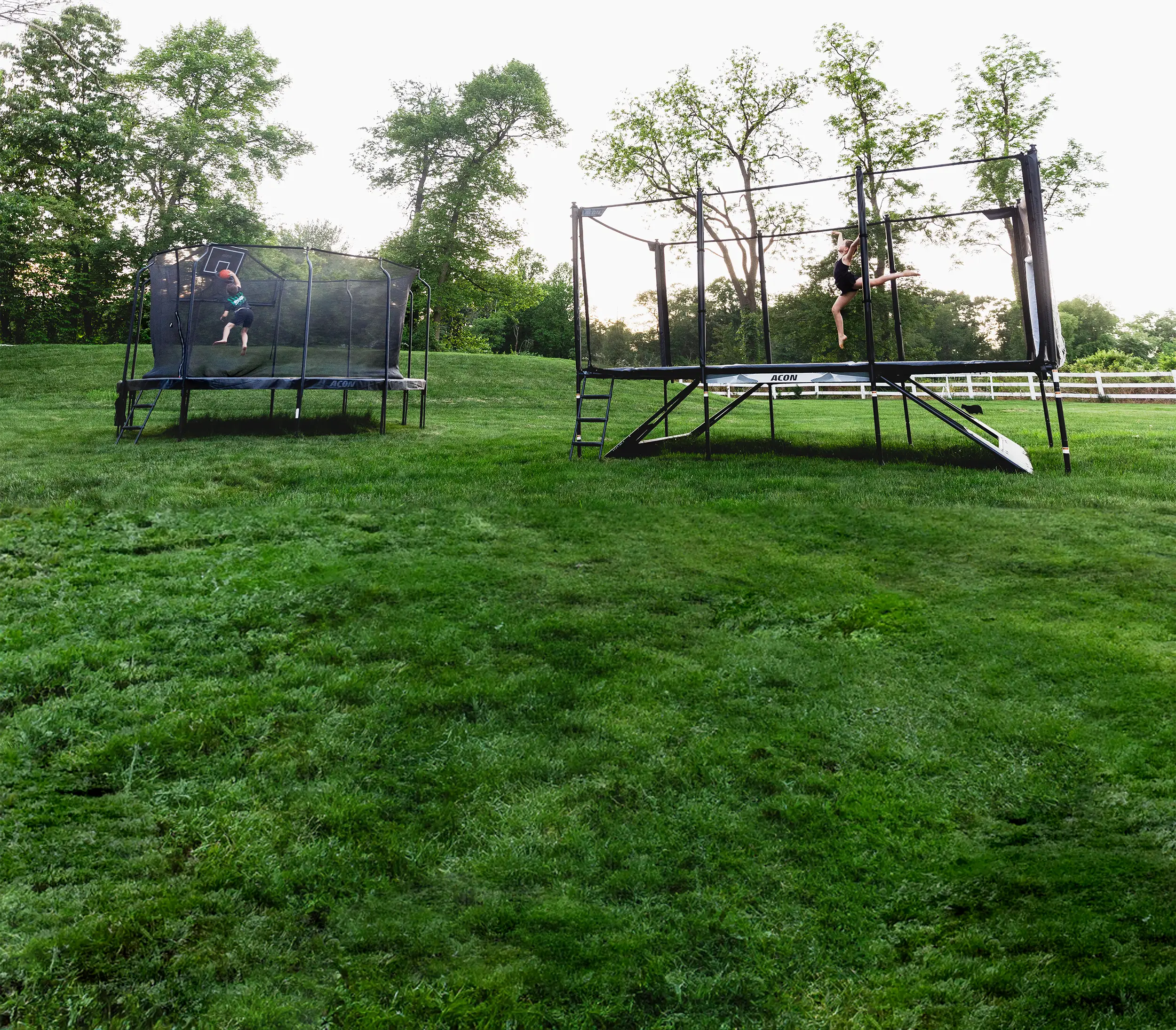 A boy is playing basketball on a round Acon trampoline, and a girl is jumping on an Acon X trampoline in a summery backyard scene.