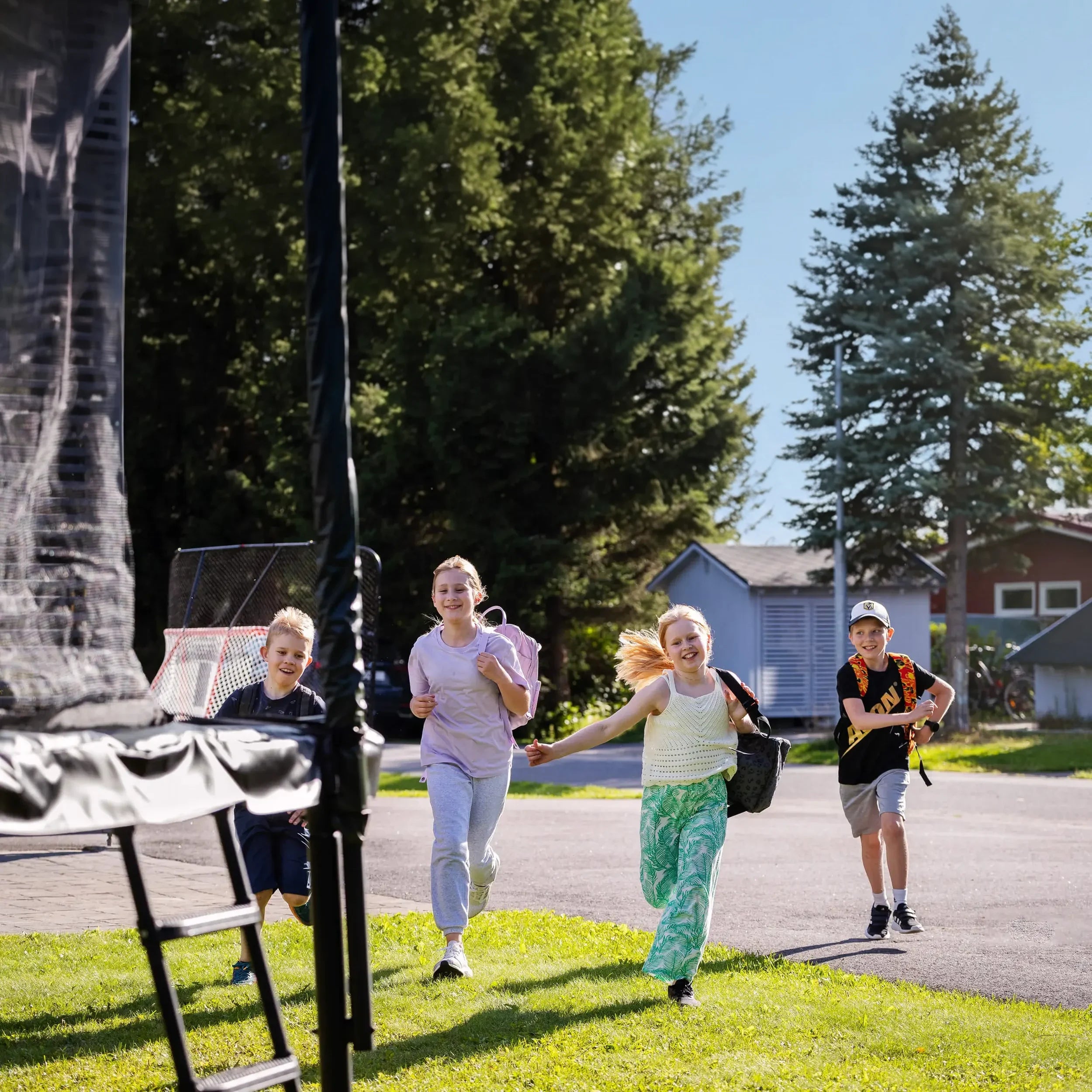 Four children run toward an Acon trampoline after school, backpacks on, in a sunny backyard.
