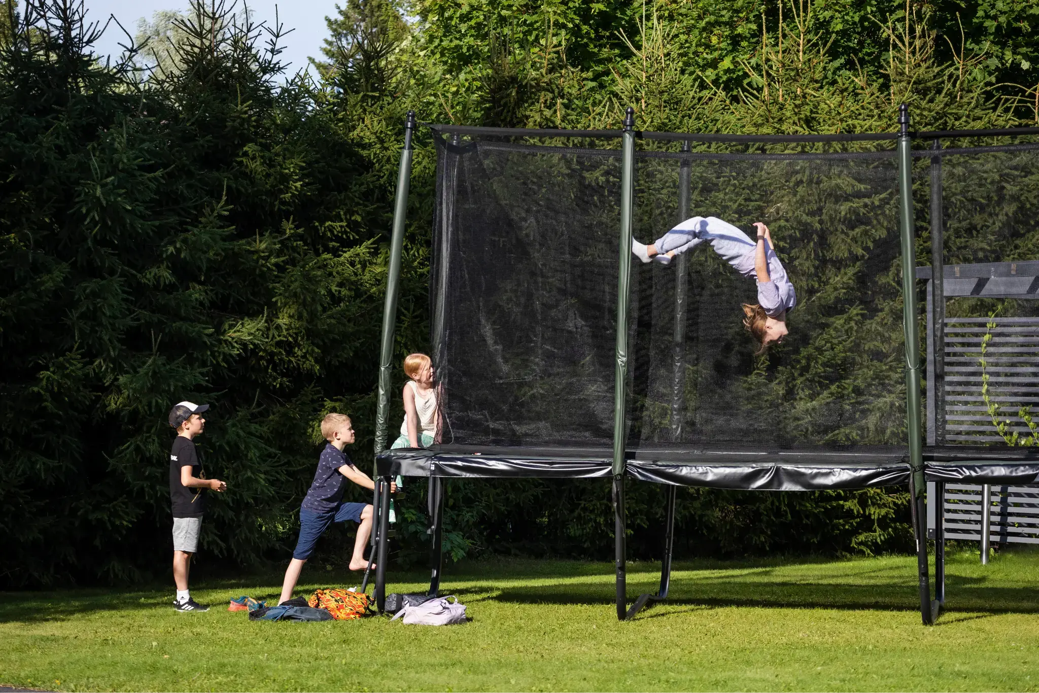 A group of children plays on an Acon trampoline in a lush backyard, with one performing a backflip while others watch and wait for their turn.