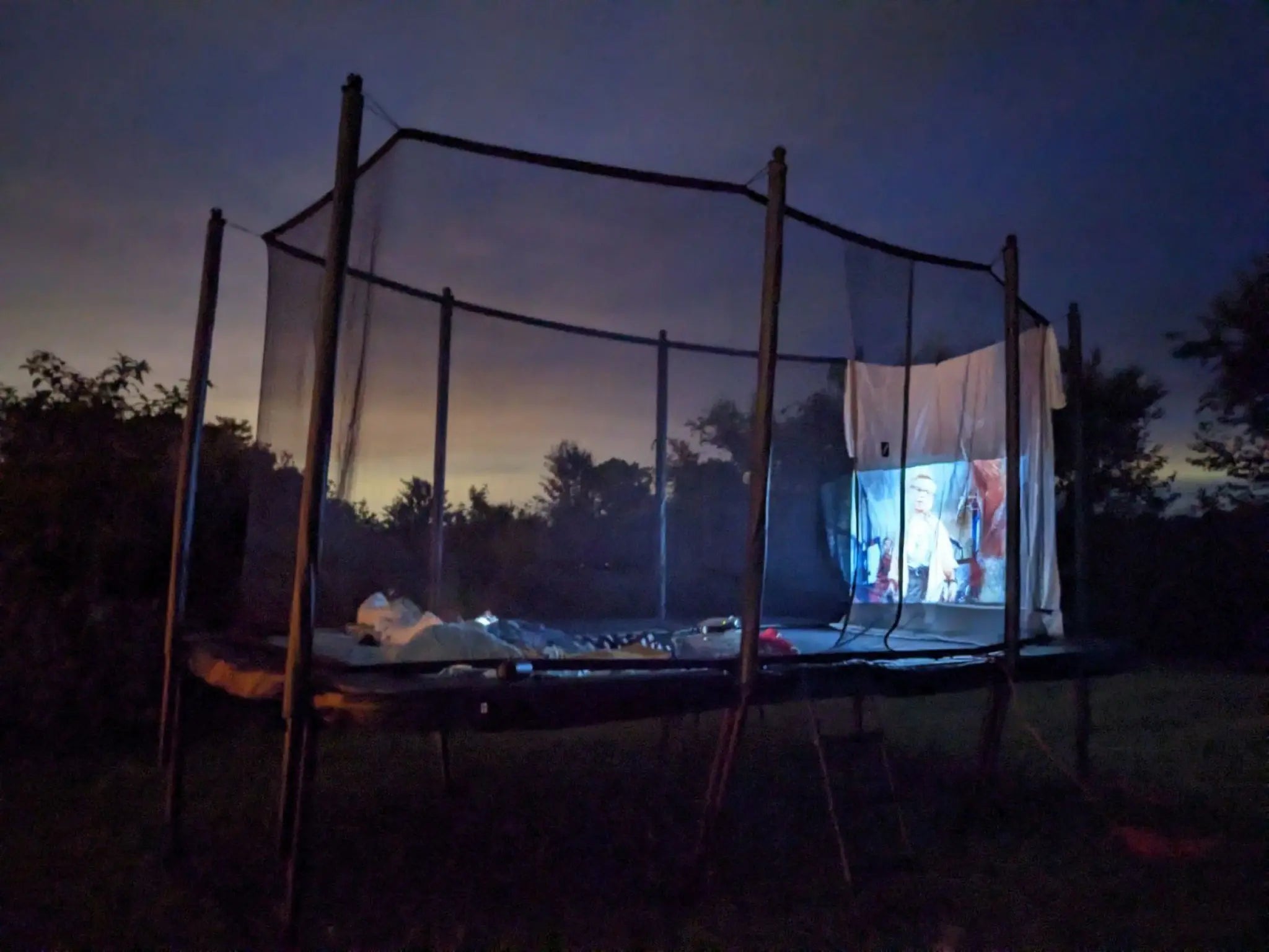 A trampoline set up with outdoor movie screen at night, with a film projected onto a white sheet and surrounded by a dark evening sky.